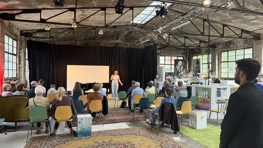 Blick von hinten auf das Publikum und die Bühne der Halle 16. Eine junge Frau hält ein Mikrofon in der Hand und moderiert den Pianisten an
