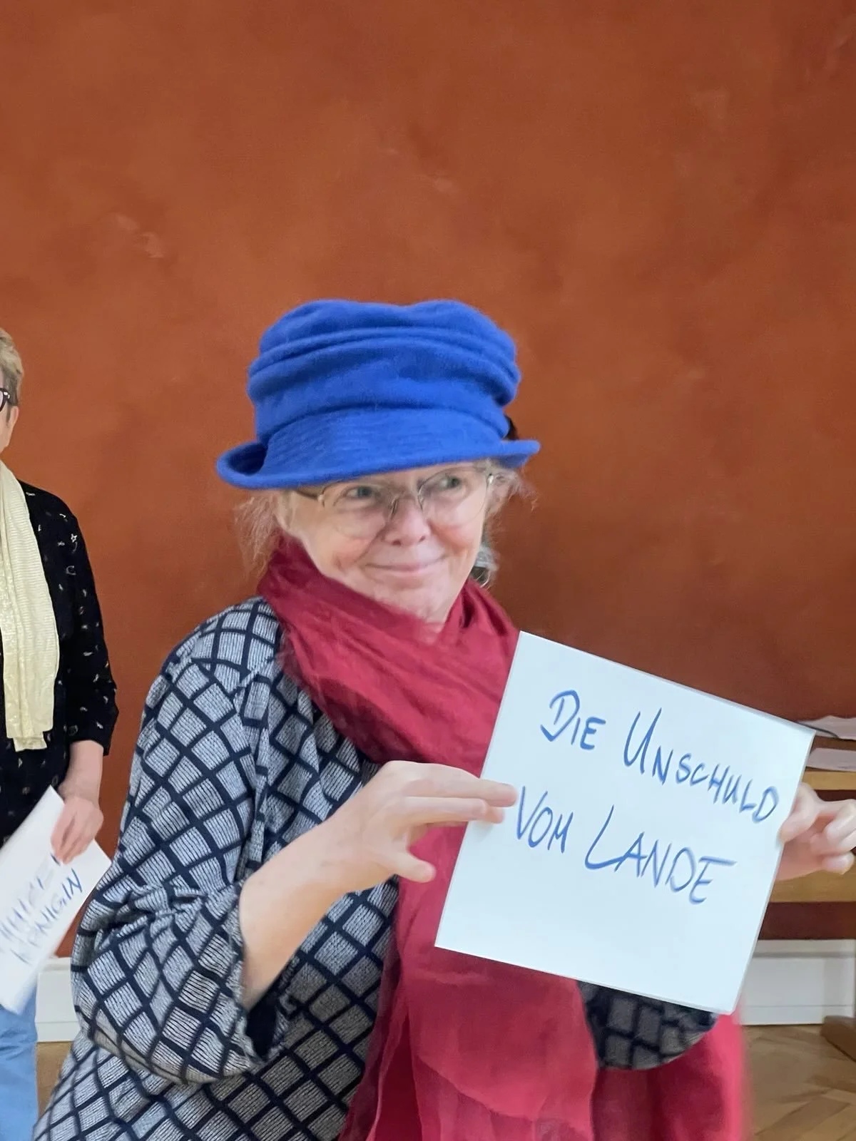 eine ältere Frau mit blauer Mütze hält schelmisch lächelnd ein  Schild in der Hand auf dem "Die Unschuld vom Lande" steht