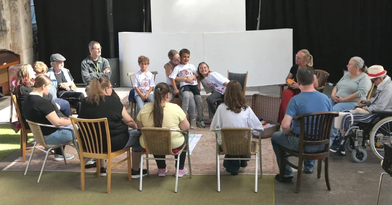 Gruppenbild der Teilnehmerinnen und Teilnehmer des Mittwochsangebotes inklusiv kreativ. Alle sitzen in einem Stuhlkreis auf den bunten Teppichen der Halle 16