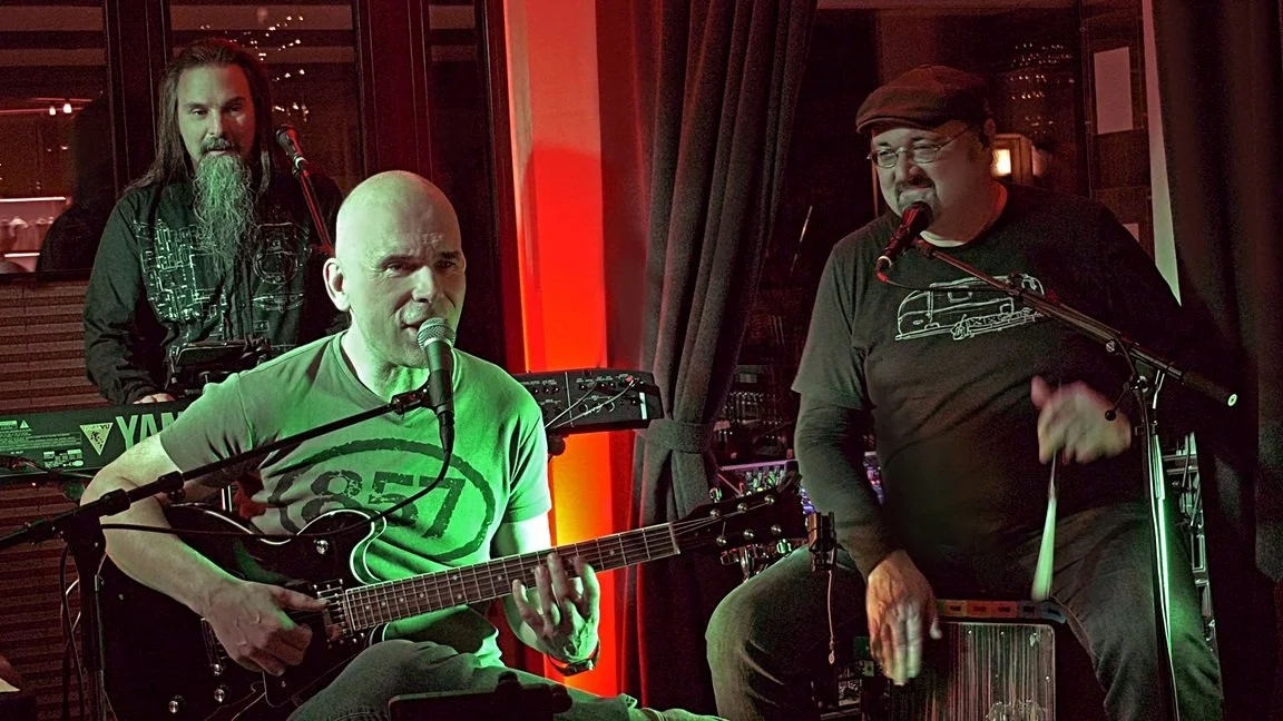 drei ältere Männer machen Musik auf einer Bühne. Ein Man mitt langem Bart spielt Keyboard im Hintergrund. Vorne in der Mitte sitzt einer mit Glatze an der E-Gitarre. Rechts außen spielt ein Mann mit Brille Percussion.