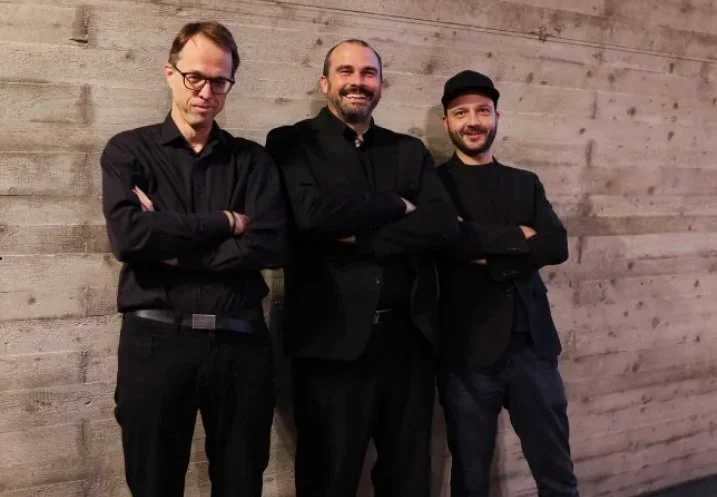 drei Männer stehen mit lächelnden und verschmitzen Gesichtsausdrücken vor einer grauen Wand