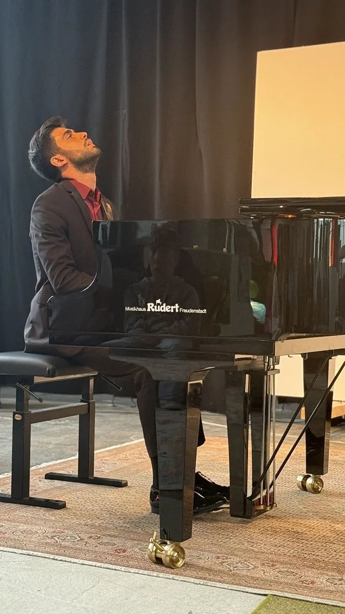 ein junger Mann in einem schwarzen Anzug sitzt an einem schwarzen Klavierflügel, ganz ins Spiel vertieft wirft er seinen Kopf in den Nacken