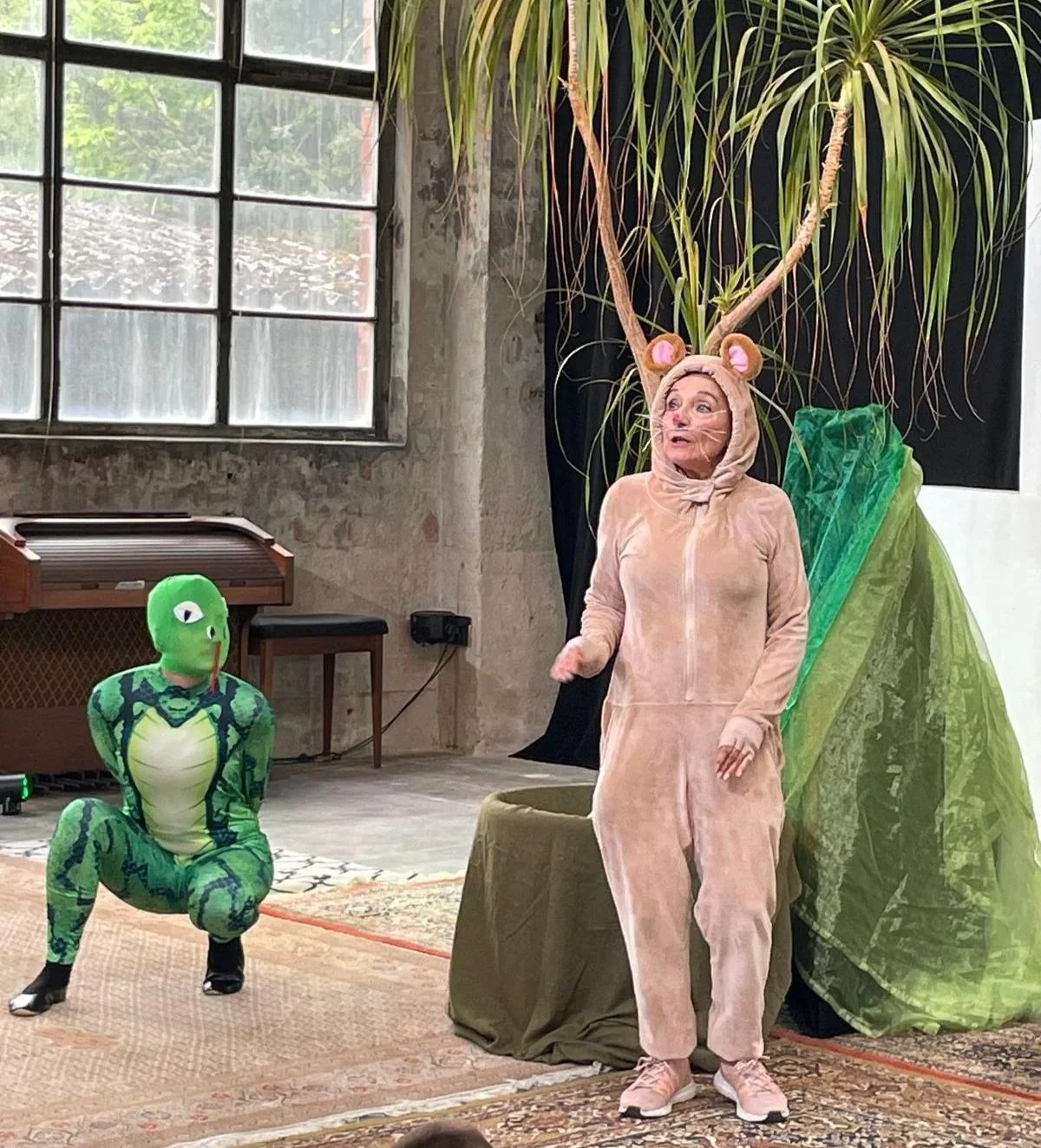In einem Wald, bestehend aus einer Palme und grünen Tüchern, steht eine als Maus verkleidete Person, sie schaut unruhig ins Publikum, neben ihr kniet eine als Schlange verkleidete Person, welche die Maus beäugt