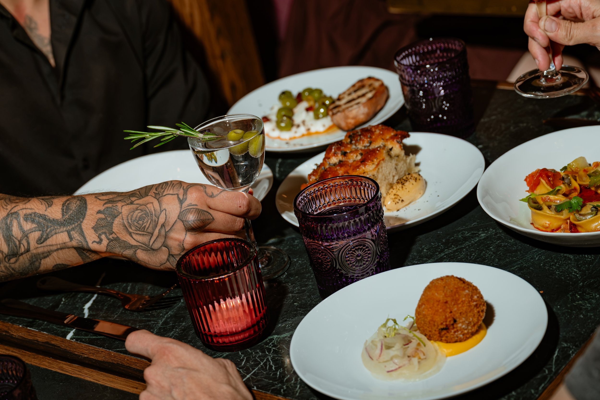 People dining at a restaurant with plates of food, glasses, and colorful glasses on the table, one person with a tattooed arm holding a small glass with olives and a sprig of rosemary.