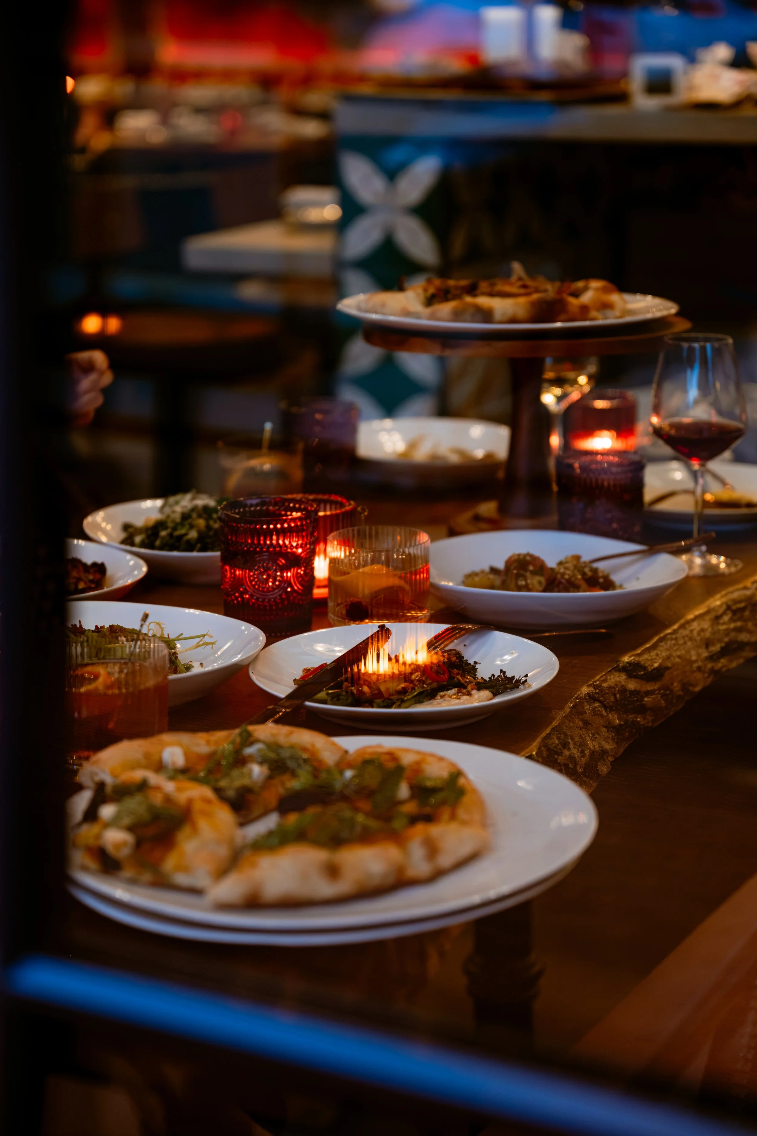 A table filled with plates of food, wine glasses, and candle holders, with a pizza on a raised platter in the foreground and other dishes and drinks in the background, in a cozy, dimly lit restaurant.