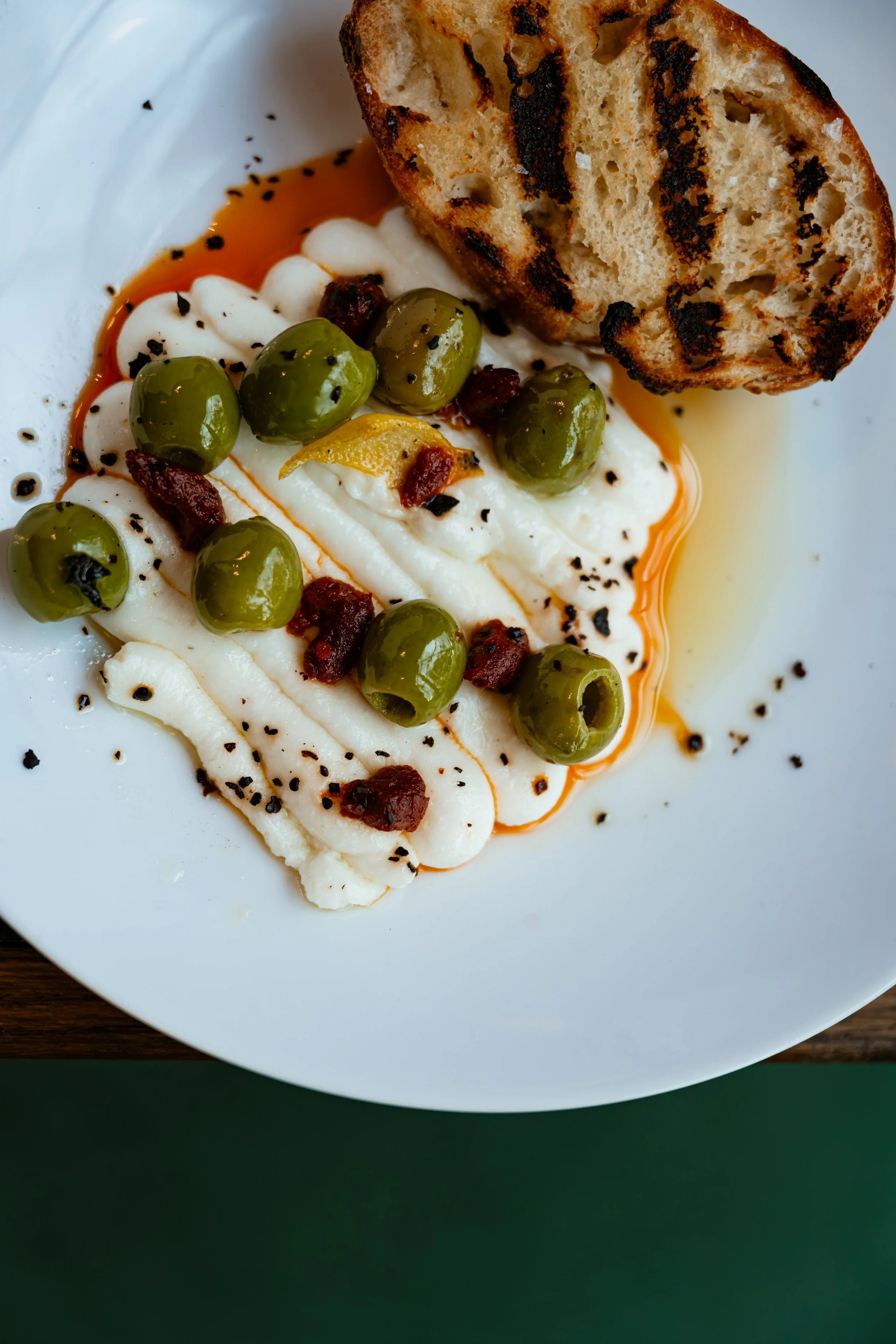 Close-up of a white plate with grilled bread, green olives, dollops of creamy spread, small bits of sun-dried tomato, black sesame seeds, and a drizzle of oil.