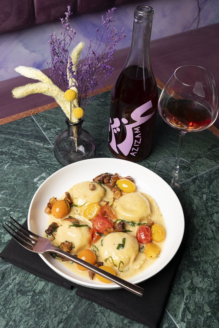Plate of ravioli pasta topped with cherry tomatoes, chopped herbs, and sauce, with a fork resting on a black napkin on a green marble table, a glass of red wine, a bottle of rosé wine, and a vase with dried purple and cream-colored flowers on the table.