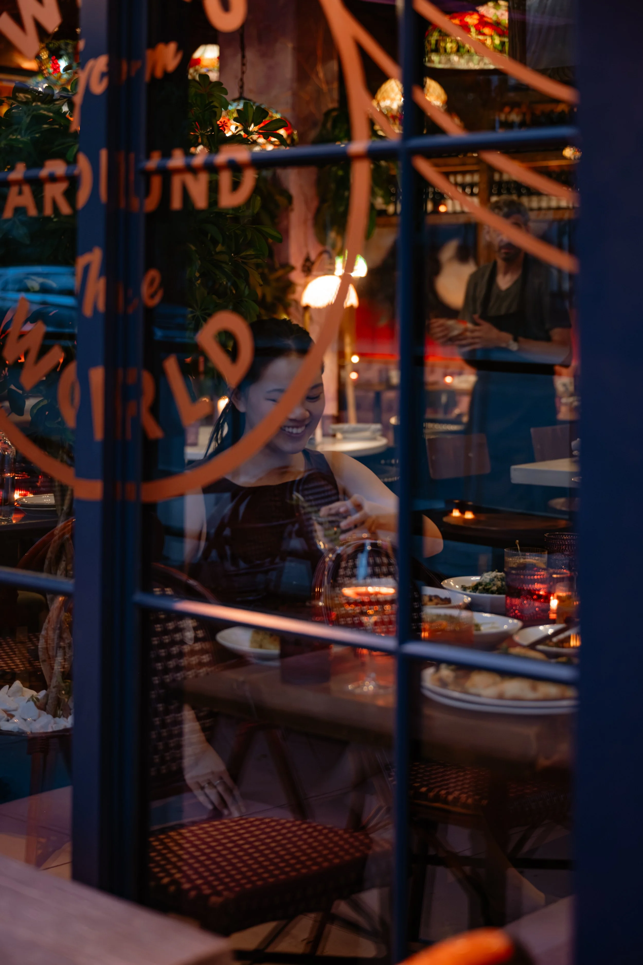 A woman sitting inside a restaurant, smiling and looking at her phone, with plates of food on the table. A server is standing in the background near colorful lighting and decorations.
