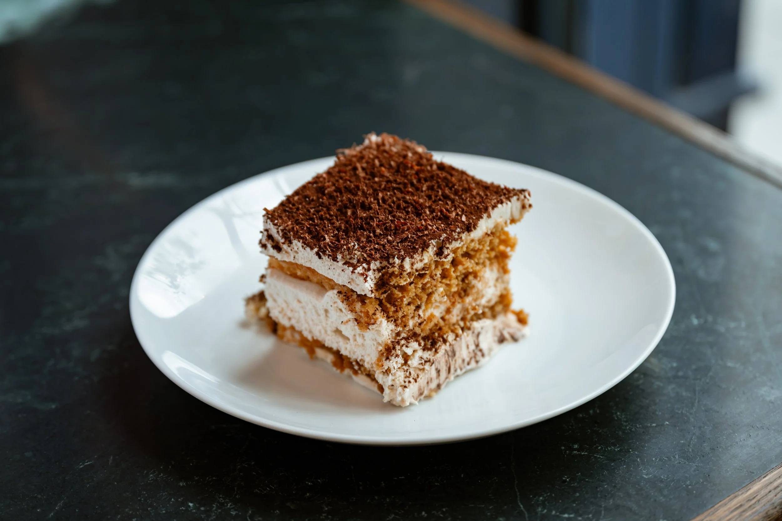 A slice of layered dessert with a crumbly base, creamy middle layer, and chocolate grated topping on a white plate.
