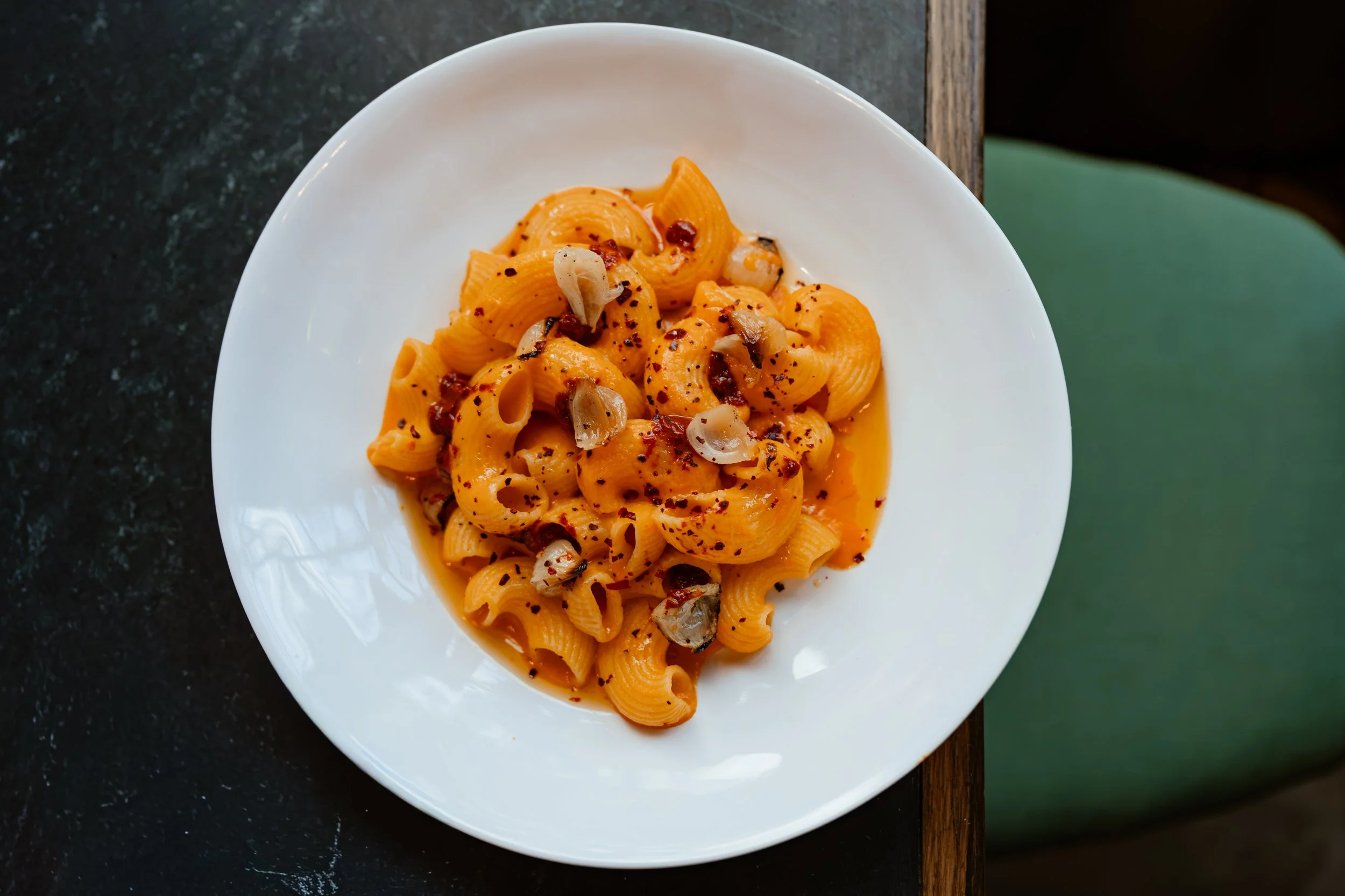 A white plate of pasta with orange sauce, garnished with chopped onions and red pepper flakes, set on a dark countertop with a green chair partially visible.
