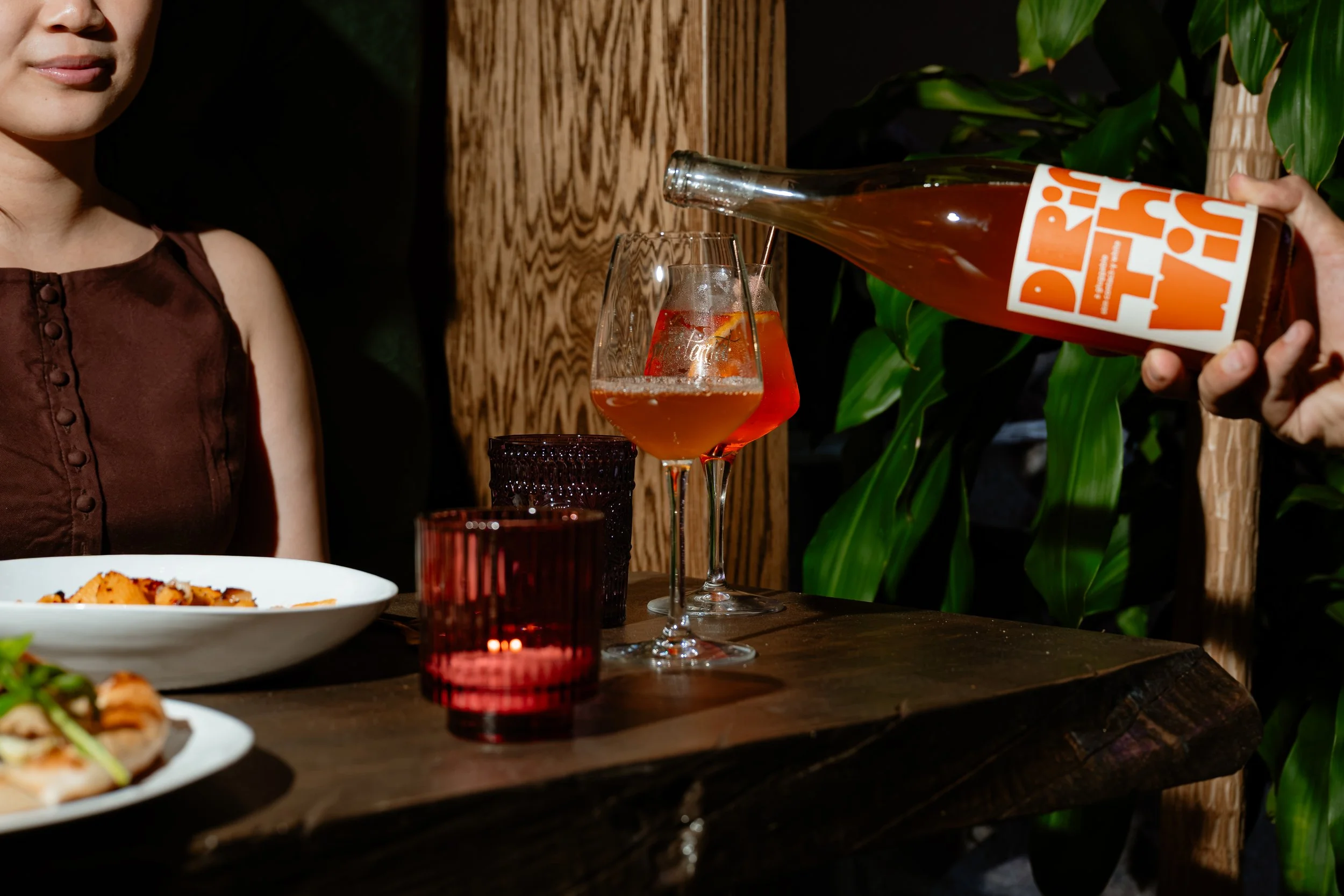 A person pouring a pinkish drink from a bottle labeled 'PERRI' into a wine glass with ice and fruit slices at a dinner table with food and drinks, surrounded by green plants and wooden decor.
