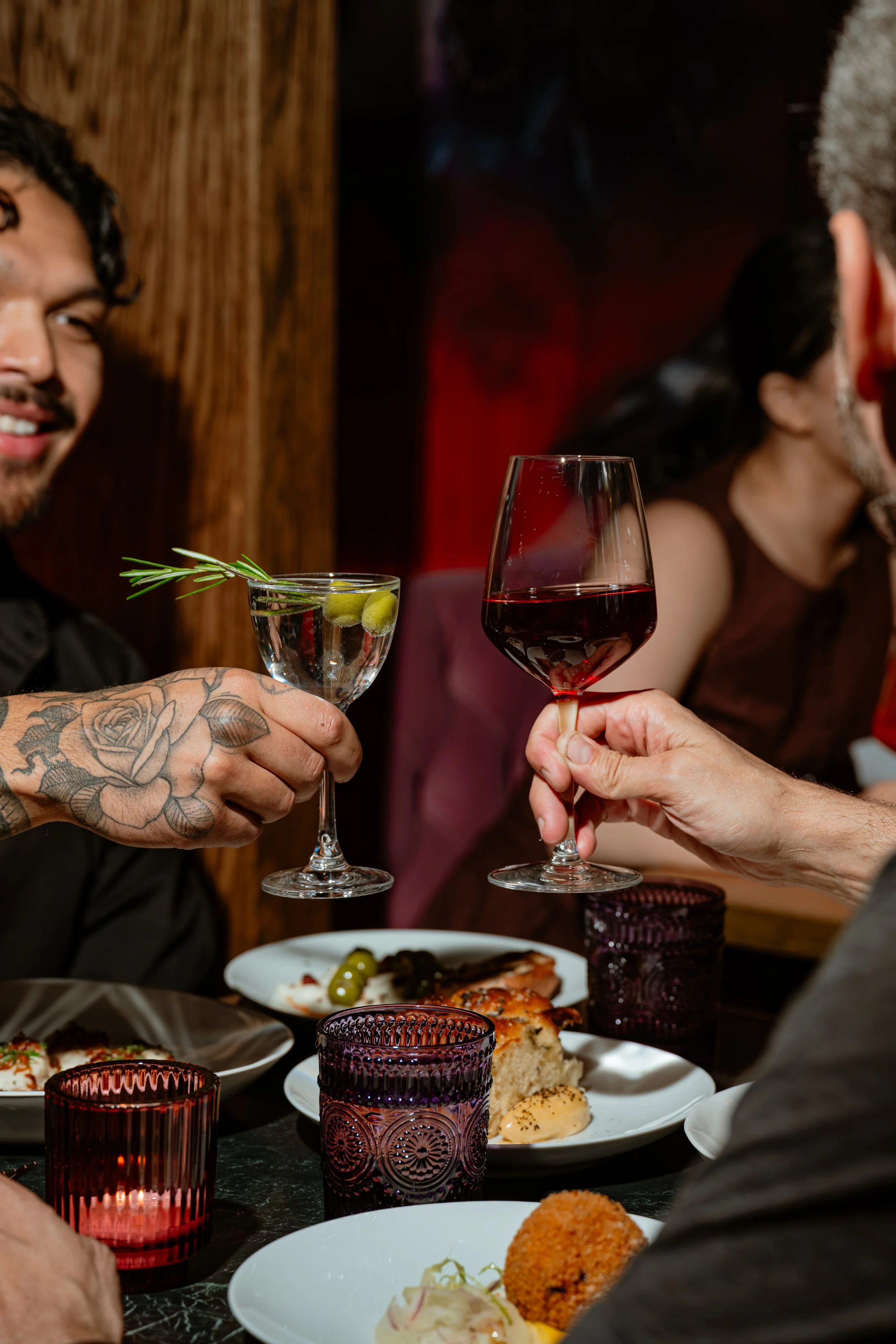 People raising glasses with drinks at a dining table filled with various dishes and lit candles.