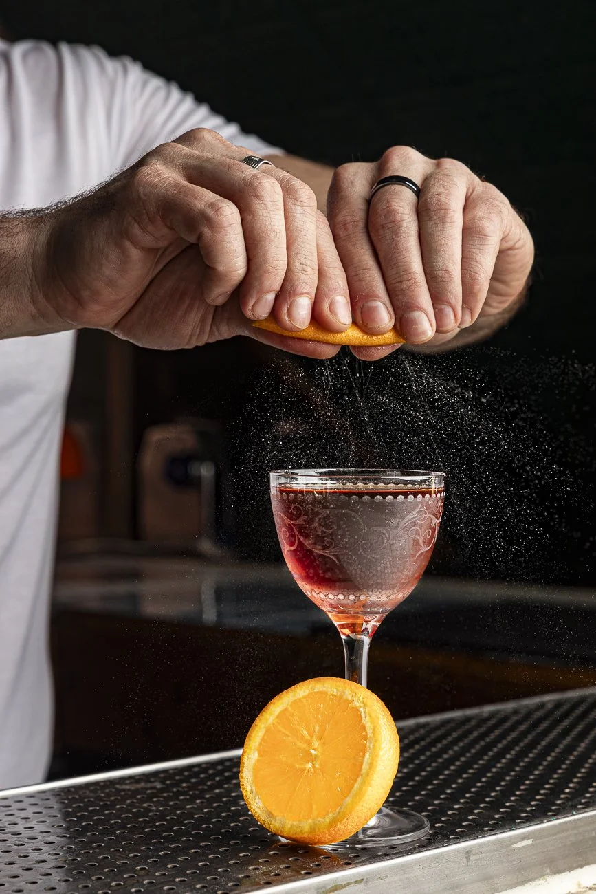 Person squeezing orange over a glass of red drink with a halved orange in front.