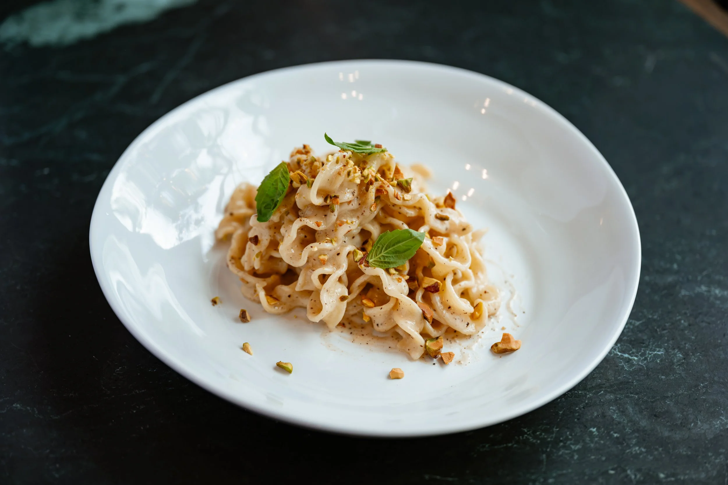 A white bowl of creamy pasta garnished with chopped nuts and fresh basil leaves, placed on a dark textured surface.