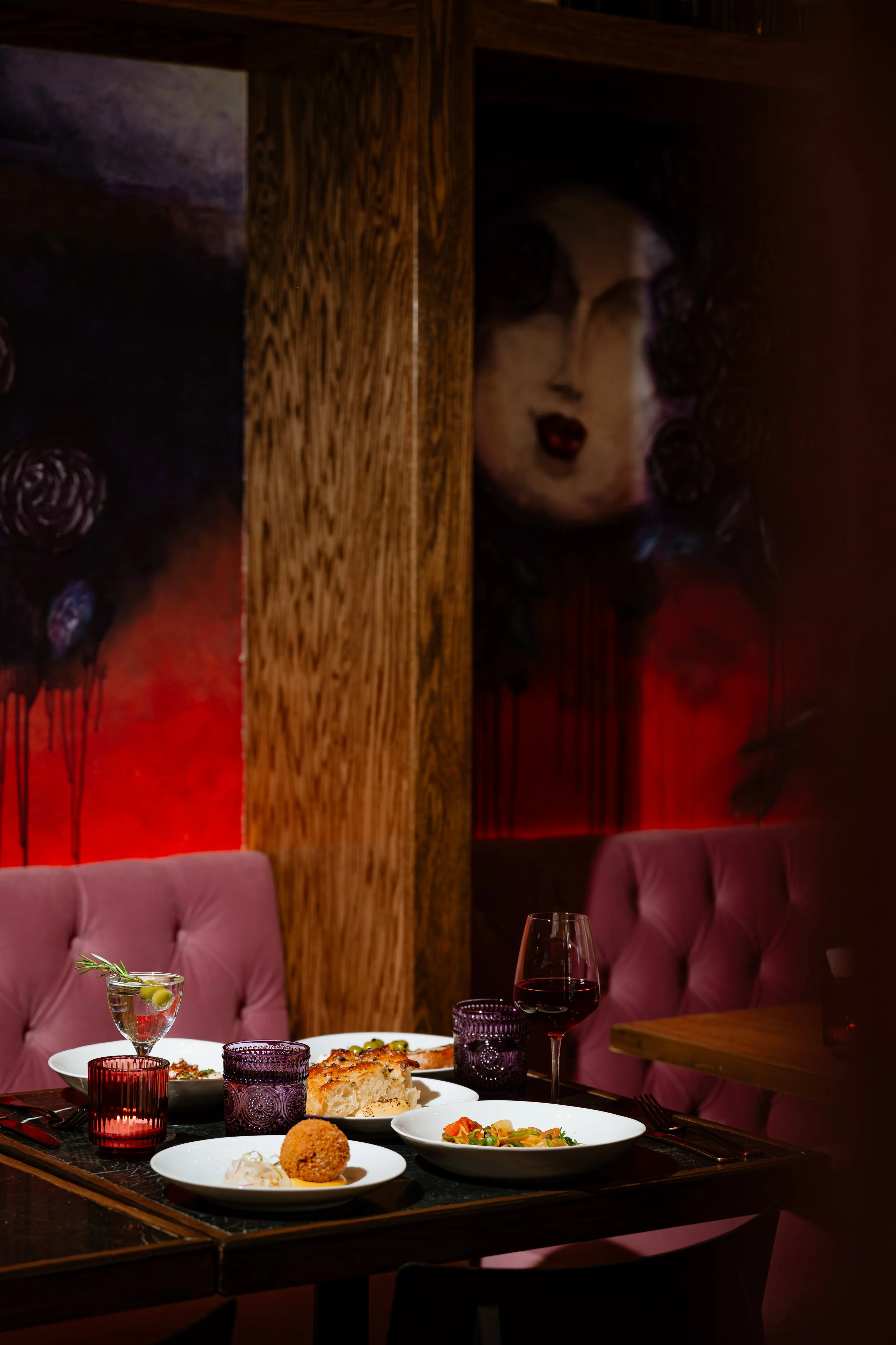 A restaurant table set with white plates of food, including a bread roll, a croquette, a salad, and a slice of pizza. There are glasses of red and white wine, and purple drinking glasses. The background has pink cushioned chairs, wooden wall partitions with artwork, and a large mural of a woman's face with dark curly hair, emphasized by red lighting.