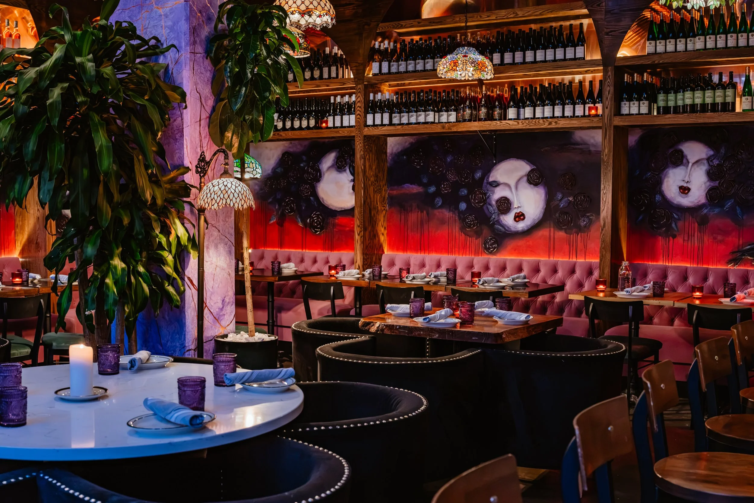 A cozy restaurant interior decorated with large paintings of female faces, wine shelves, greenery, colorful stained glass lamps, and a table set for dining with candles and purple glasses.