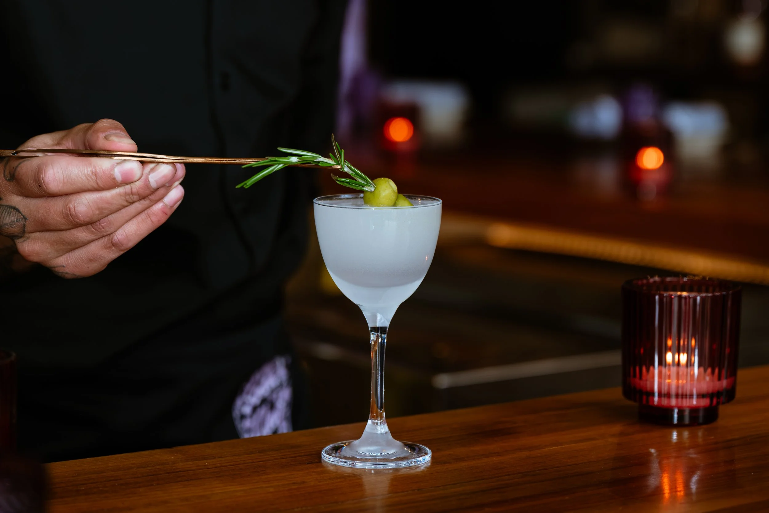 A person with tattooed fingers holding a pair of tweezers placing a sprig of rosemary and green olives into a stemmed cocktail glass filled with a clear liquid.
