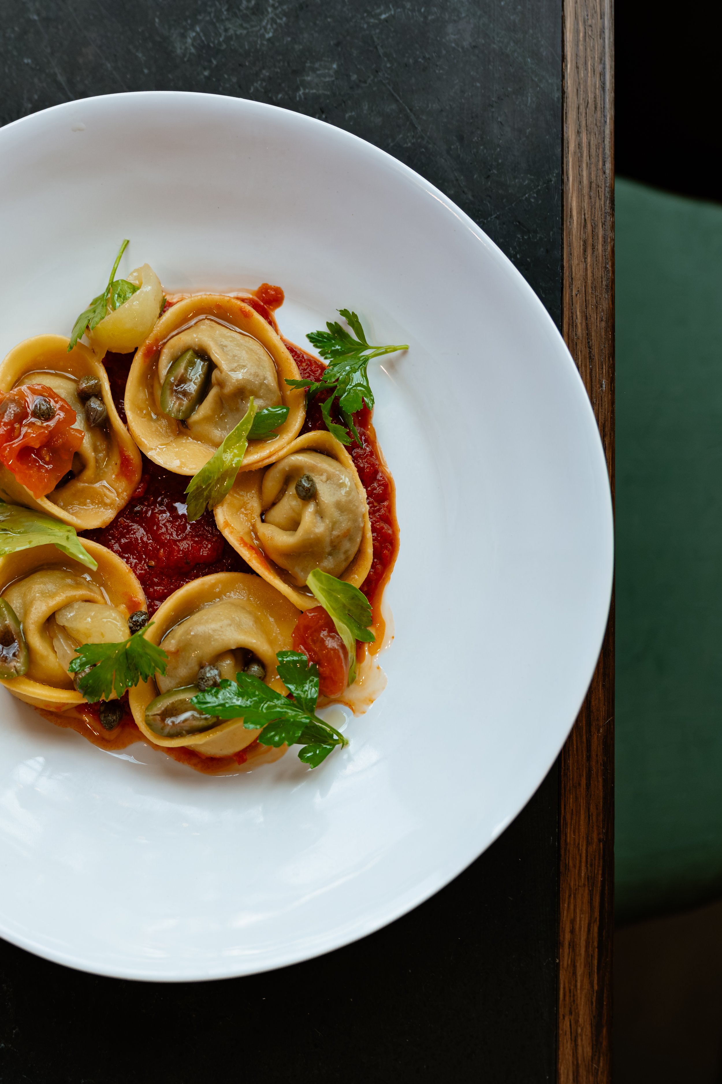 A white bowl of tortellini pasta with tomato sauce, garnished with parsley and capers, on a dark table.