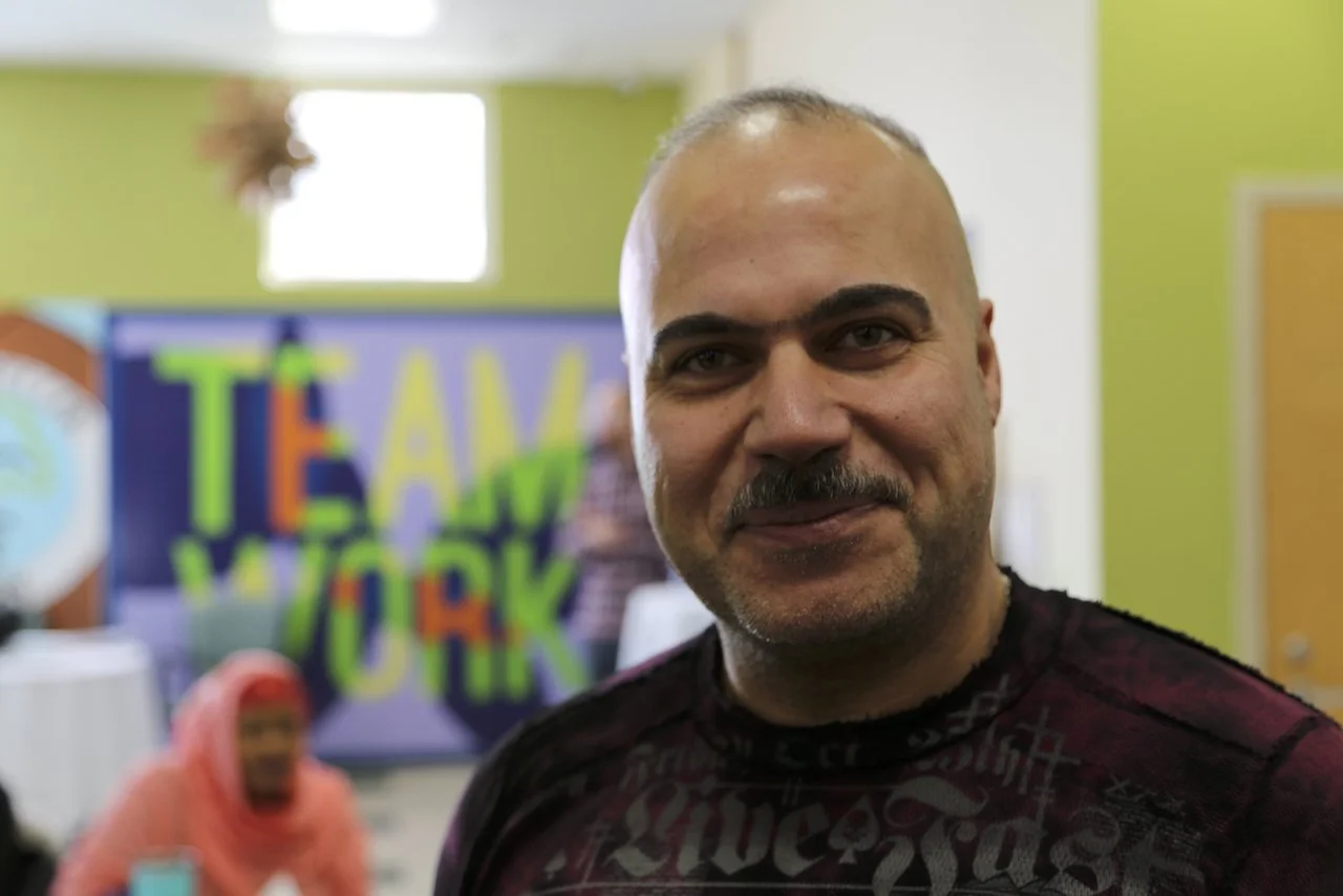 Smiling man indoors with 'Team Work' sign and people in the background.