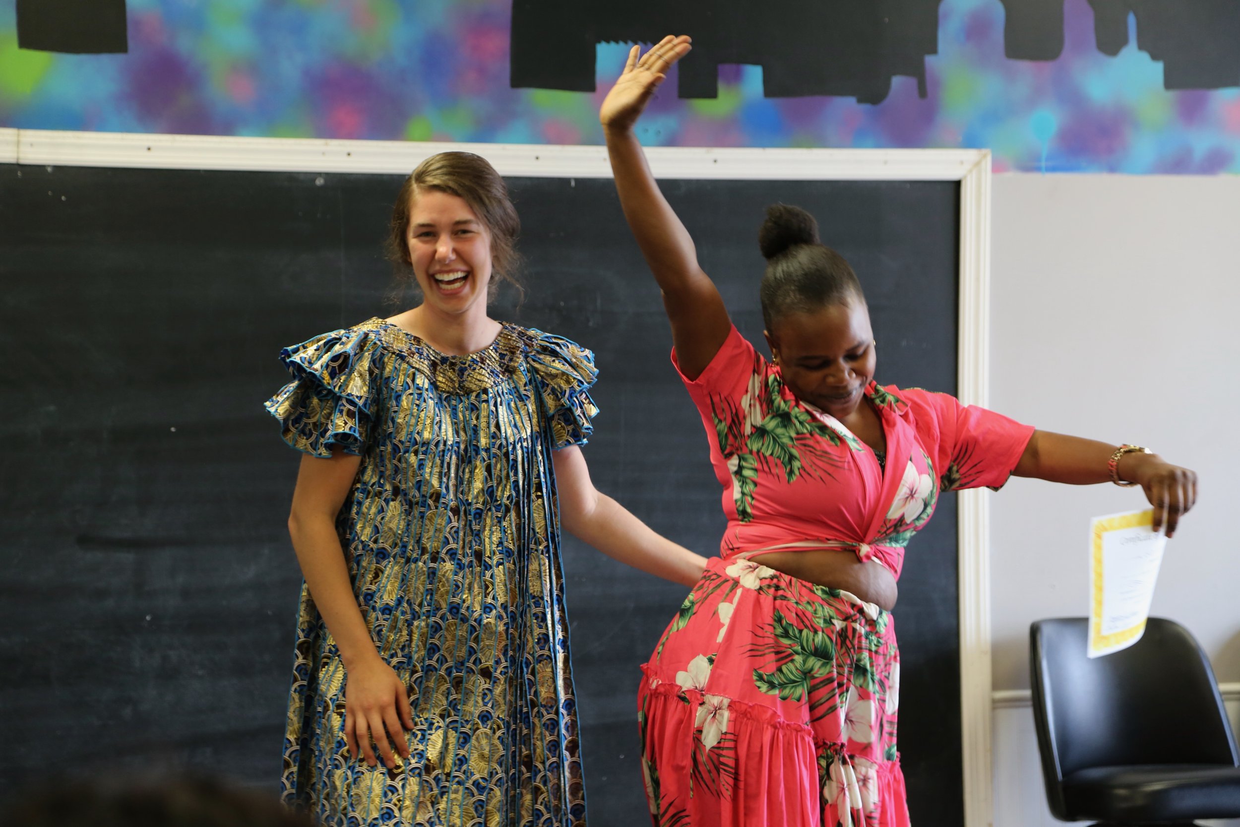 Two women in colorful dresses joyfully performing in front of a blackboard, one holding a certificate.