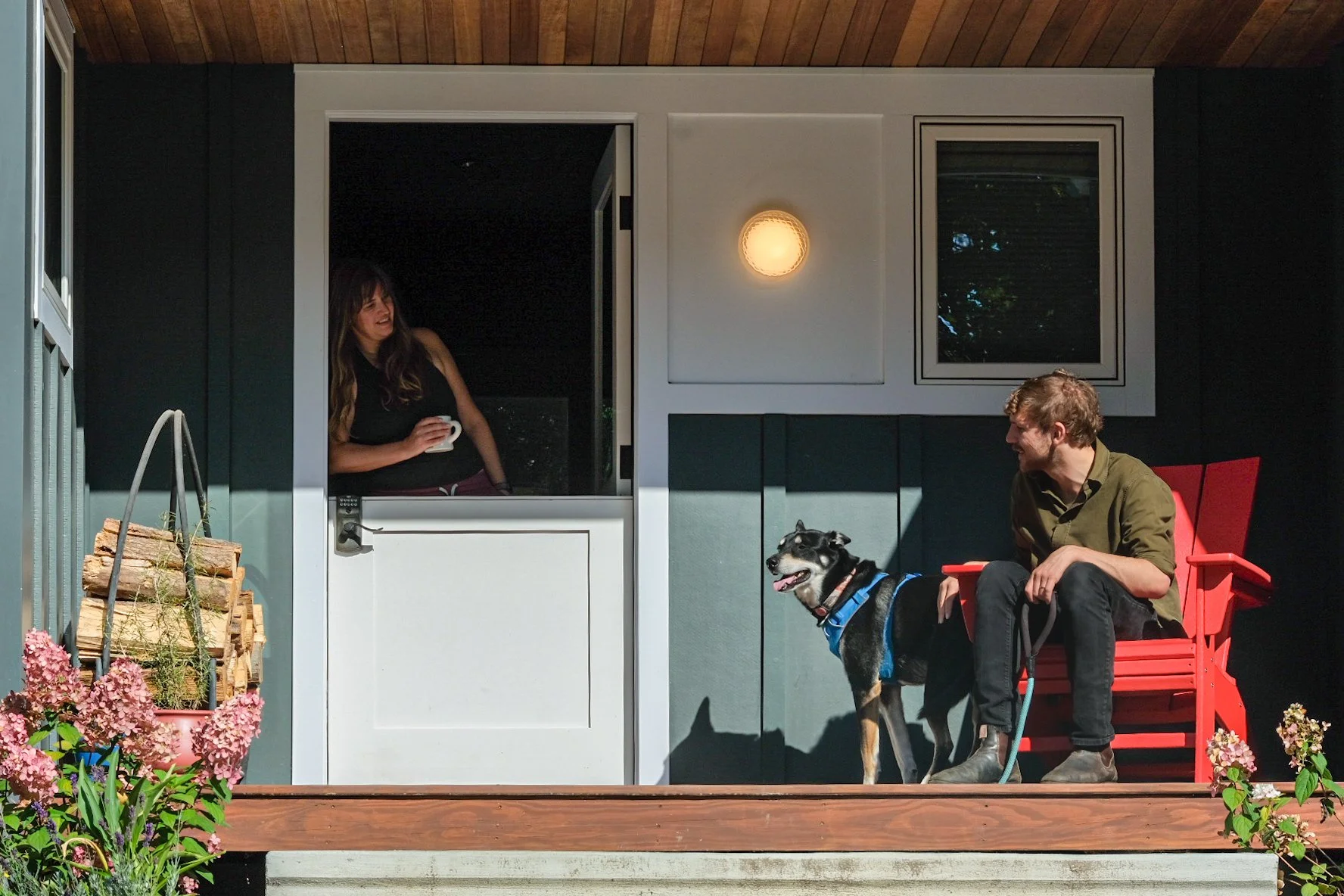 A man sitting on a red chair on a porch, holding a dog leash with a black and white dog, and a woman smiling inside the house holding a mug.