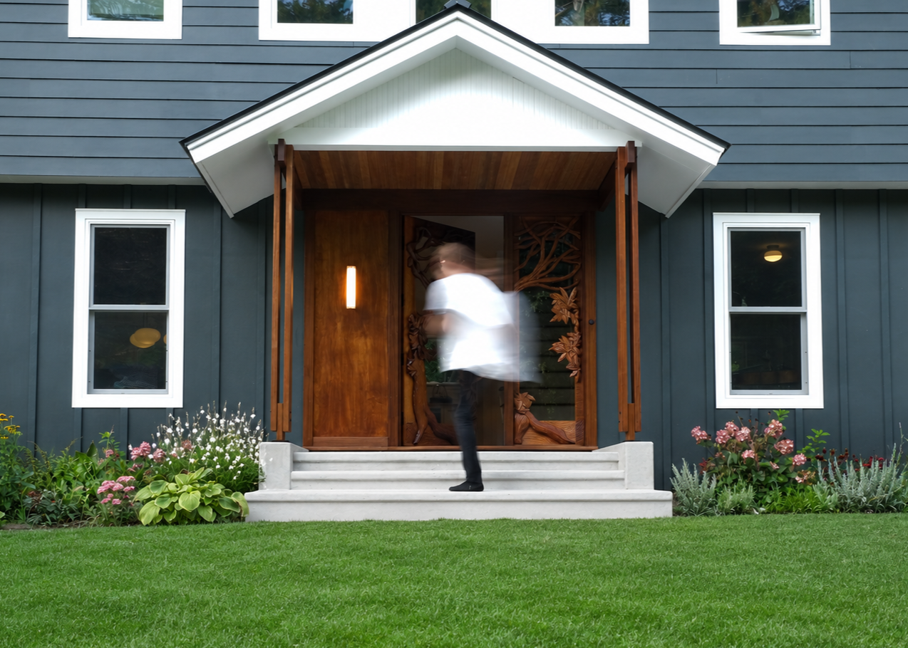 A person walking up the steps to a modern house with a blue exterior and white window frames, and a wooden carved front door. There is a lawn and flower bed in front of the house.