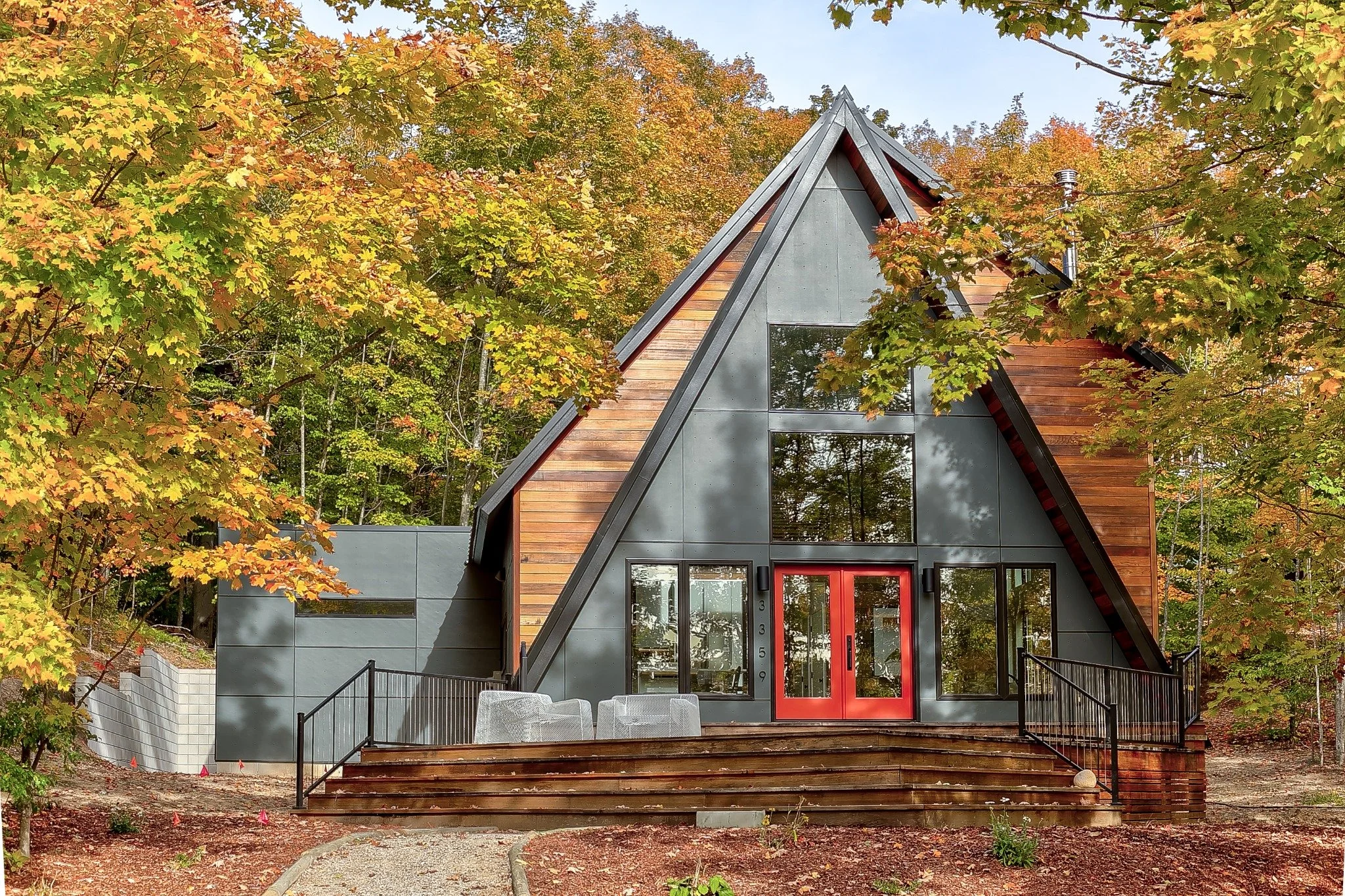  Contemporary lake house design with a steep roof, wood accents, red doors, and a stepped deck among autumn trees.
