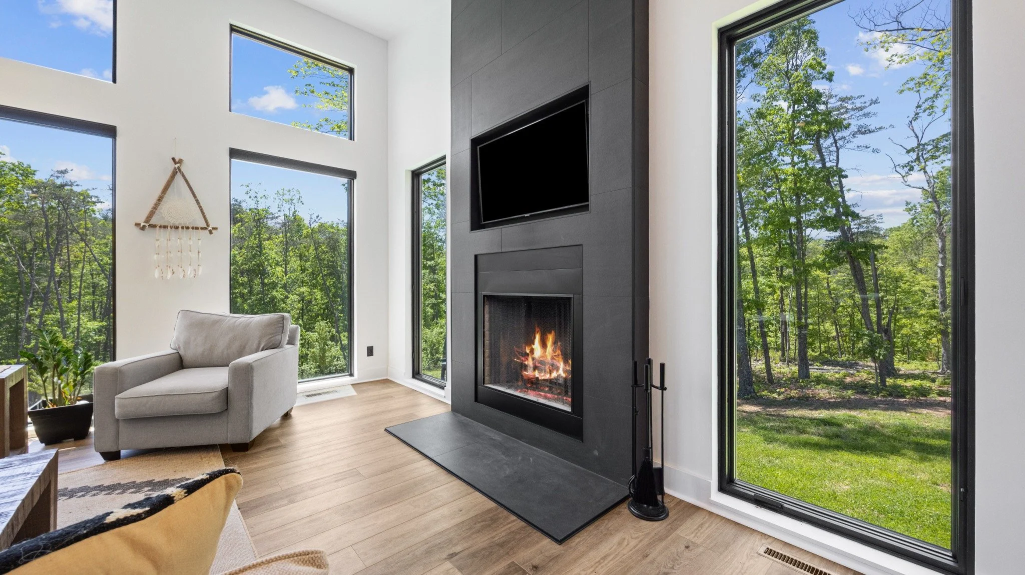 Residential architect project featuring a gray fireplace surround, upholstered armchair, wood flooring, and tall windows overlooking woodland.