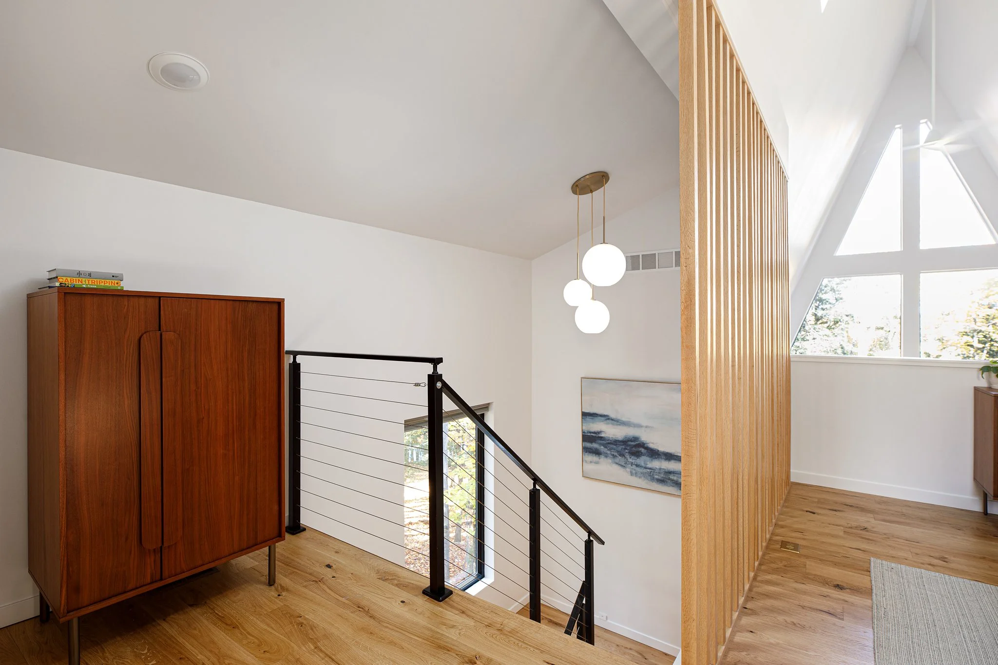 Modern home's staircase area with wooden flooring, white walls, and a black railing. A wooden cabinet with books on top and a large geometric window letting in natural light. A wooden slat room divider separates spaces, and a white pendant light hang