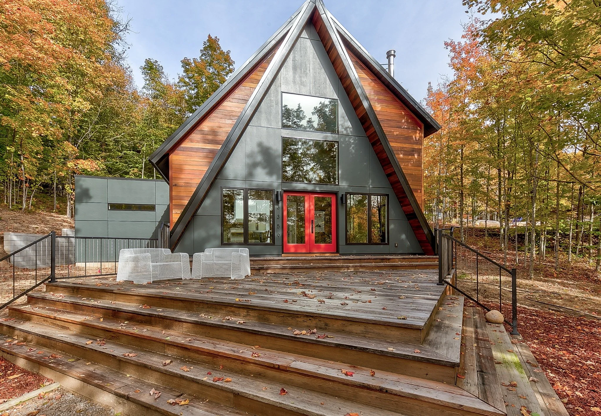 Lakeside home design with wood siding, gray panels, red double doors, and white chairs on a wood deck.