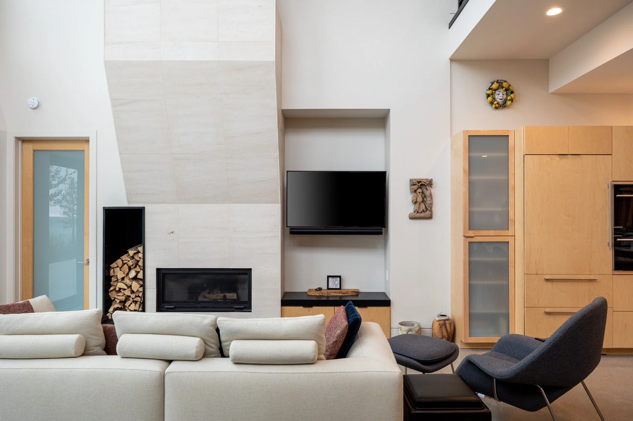 Lake house interior design with a white sofa, pale fireplace surround, firewood storage, dark lounge chair, and wood cabinetry.
