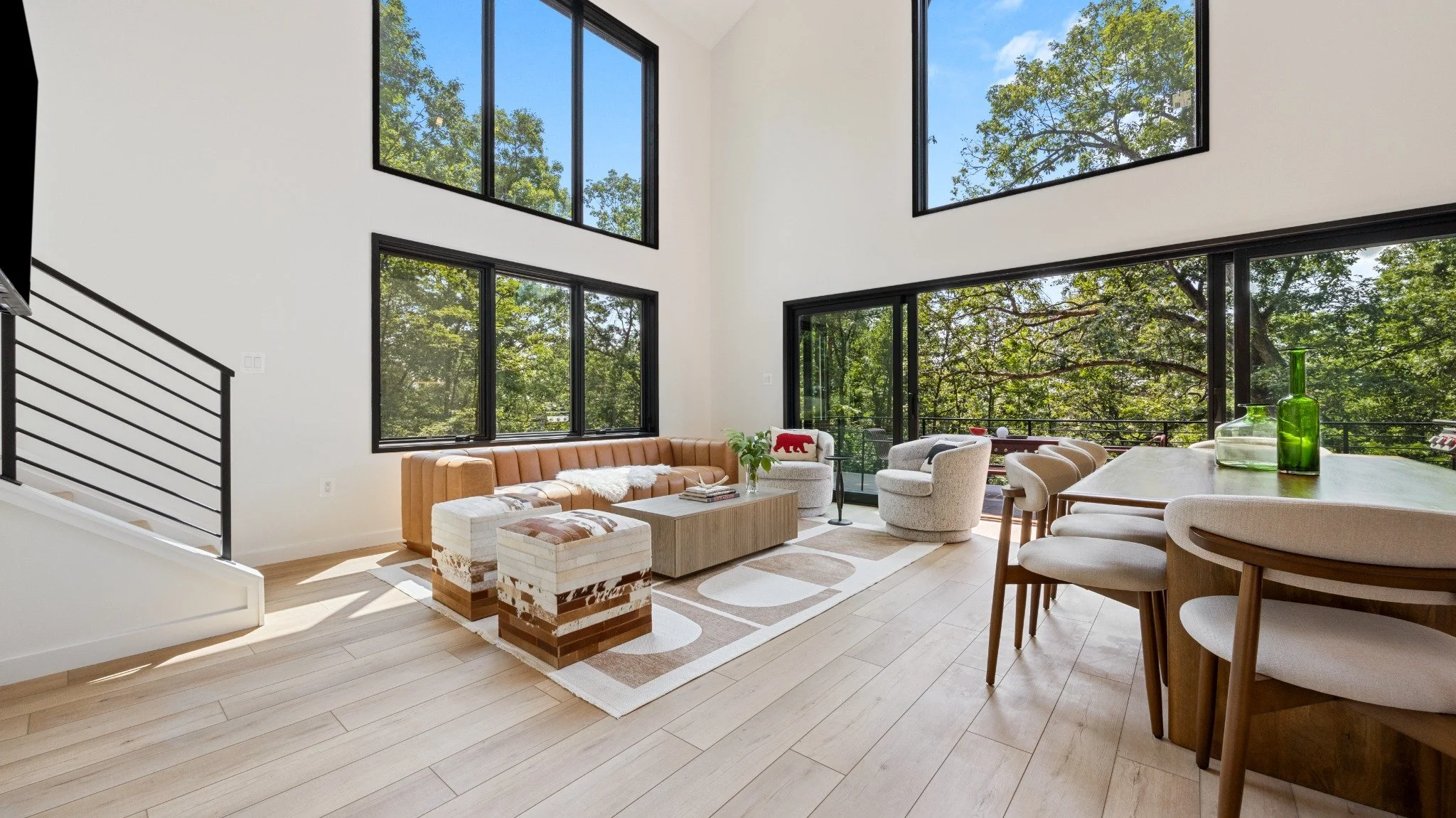  Custom mountain home architect project featuring a tan sofa, pale lounge chairs, a wood dining table, and double-height windows overlooking trees.