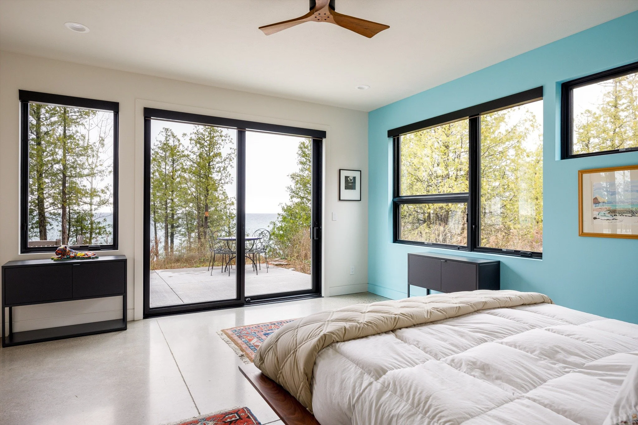  Lake house bedroom design with a blue accent wall, black-framed windows, and sliding doors to a patio overlooking water.