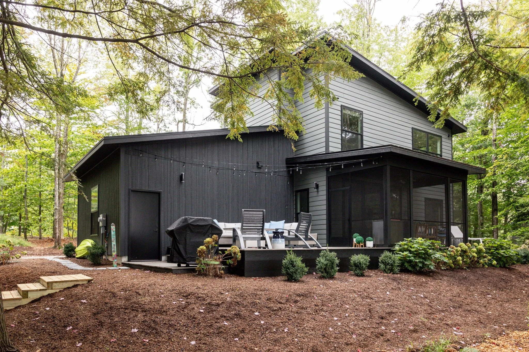Custom lake house design with mixed gray and black siding, a screened porch, and a deck with seating and string lights.

