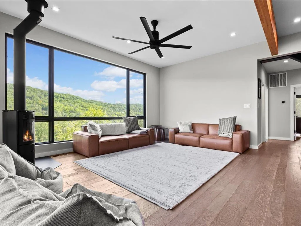Mountain house architect project featuring brown leather sofas, a black wood stove, wood flooring, and large windows overlooking wooded hills.