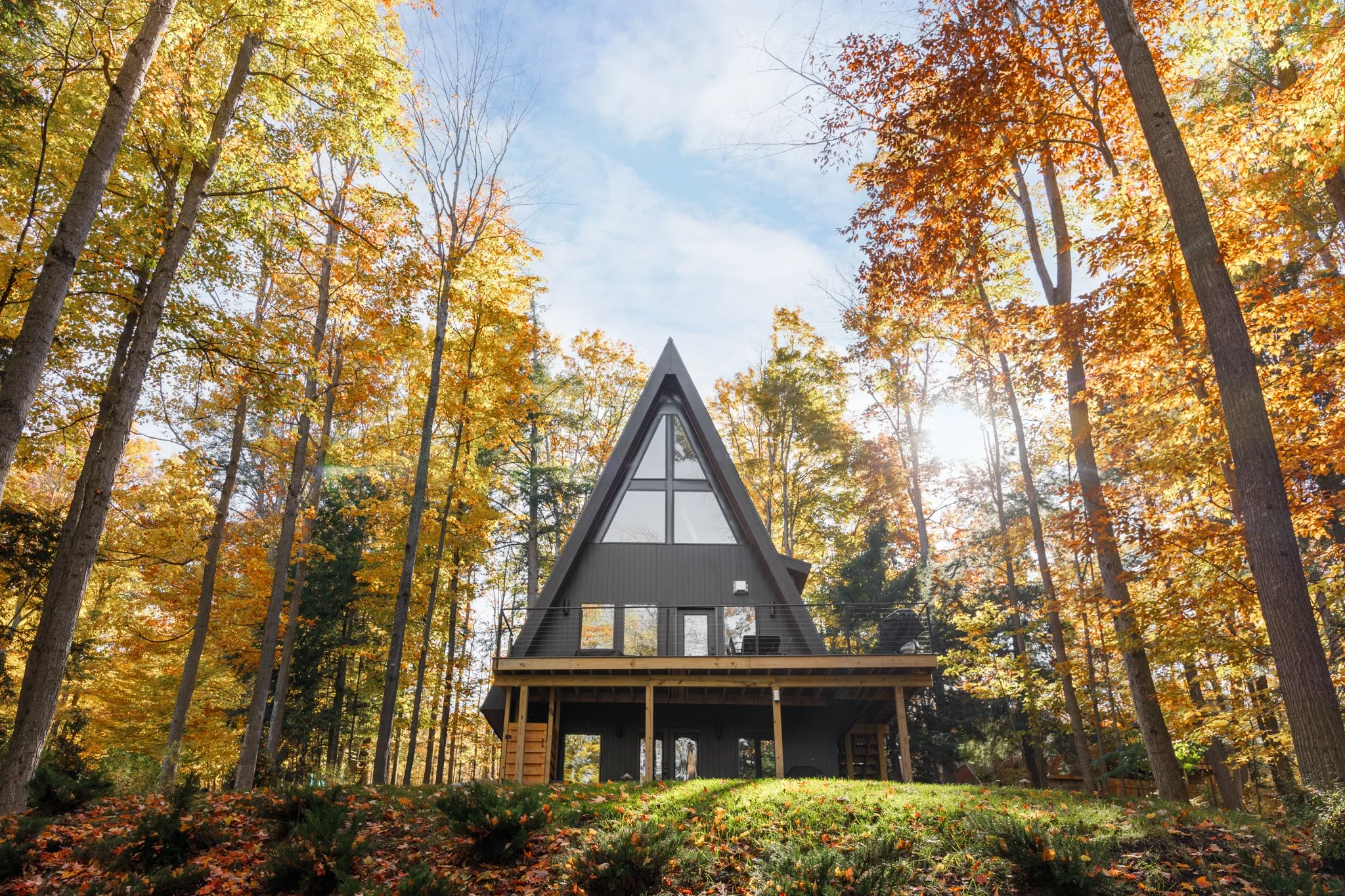 Aerial view of a modern A-frame house in a forest during autumn, with trees displaying yellow and orange leaves, and the house having a deck and large windows.