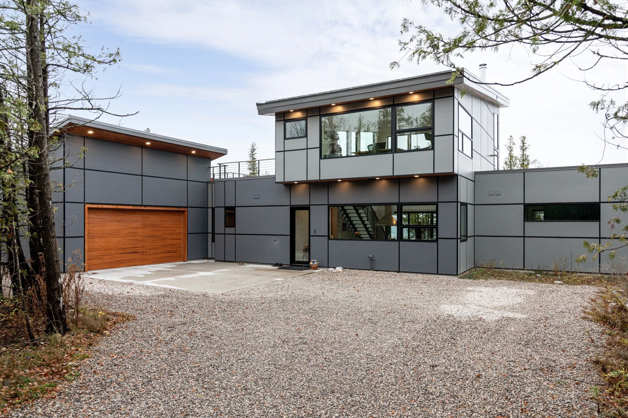Custom lake house design with gray panel siding, a wood garage door, large windows, and a gravel driveway.
