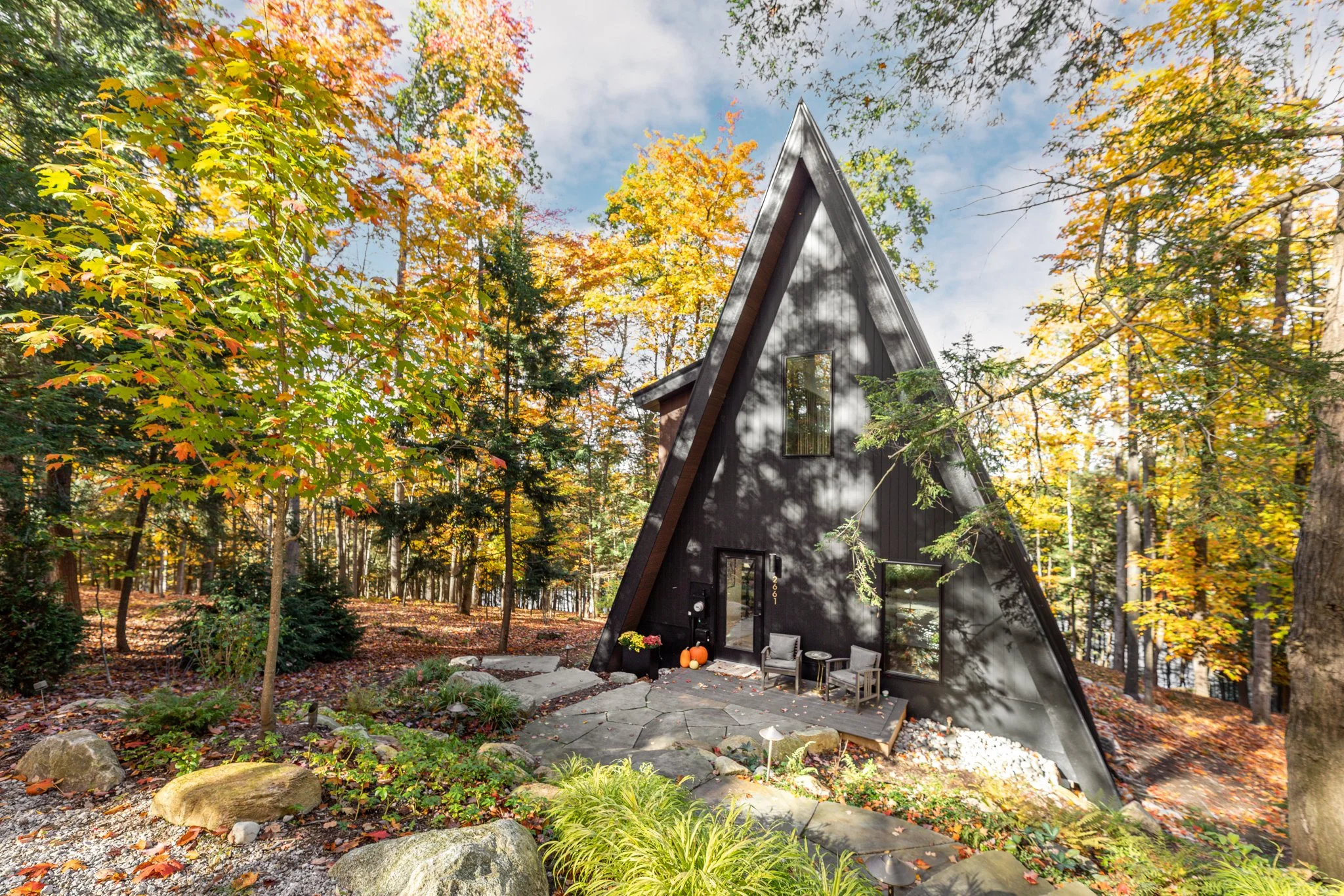 Black A-frame cabin surrounded by autumn trees and foliage, with a small outdoor seating area and pumpkins near the door.