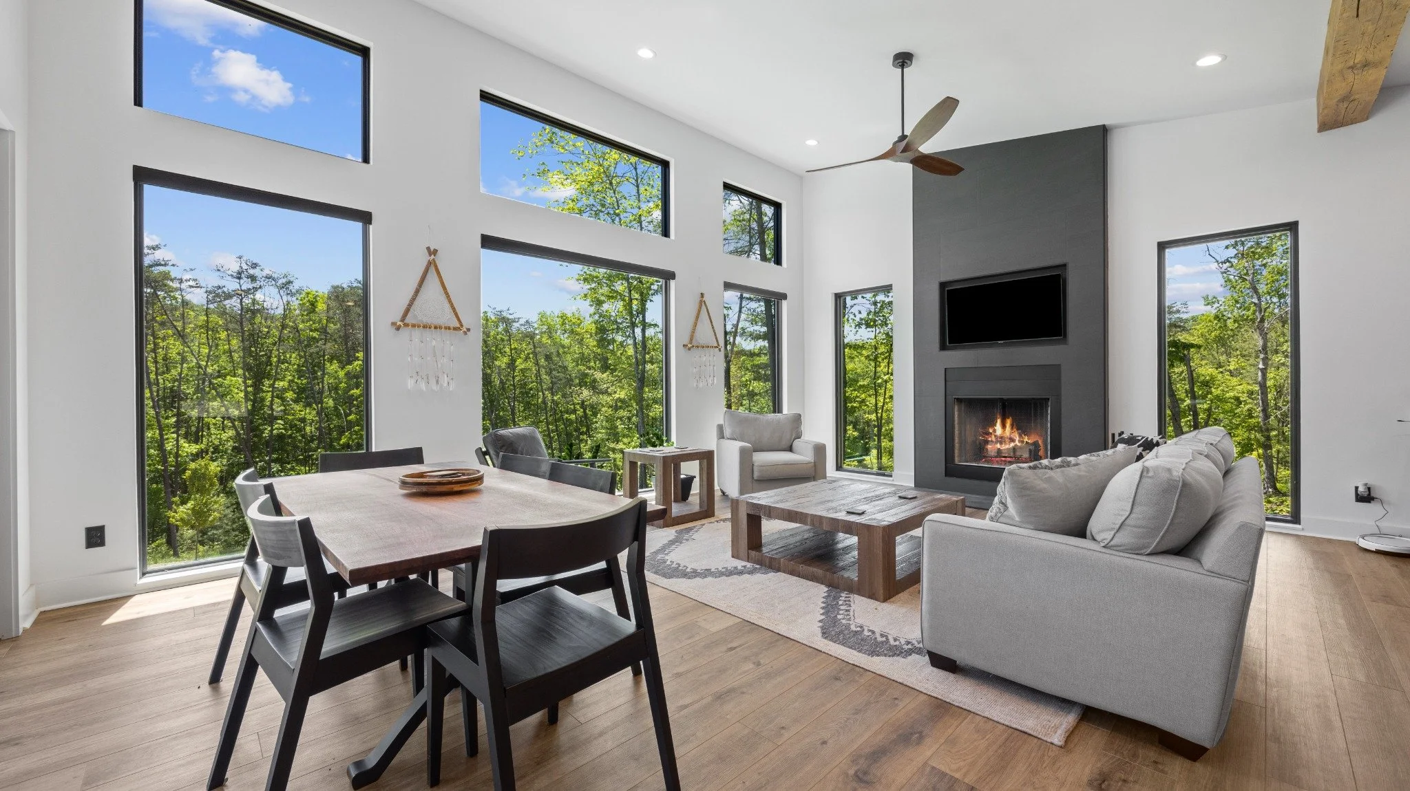 Modern mountain home architect project featuring a gray sofa, wood dining table, tall gray fireplace surround, and large windows overlooking trees.