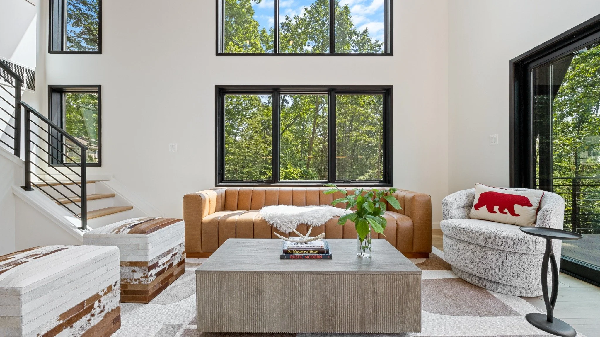 Residential architect project featuring a tan sofa, pale lounge chair, patterned ottomans, and tall black-framed windows overlooking trees.
