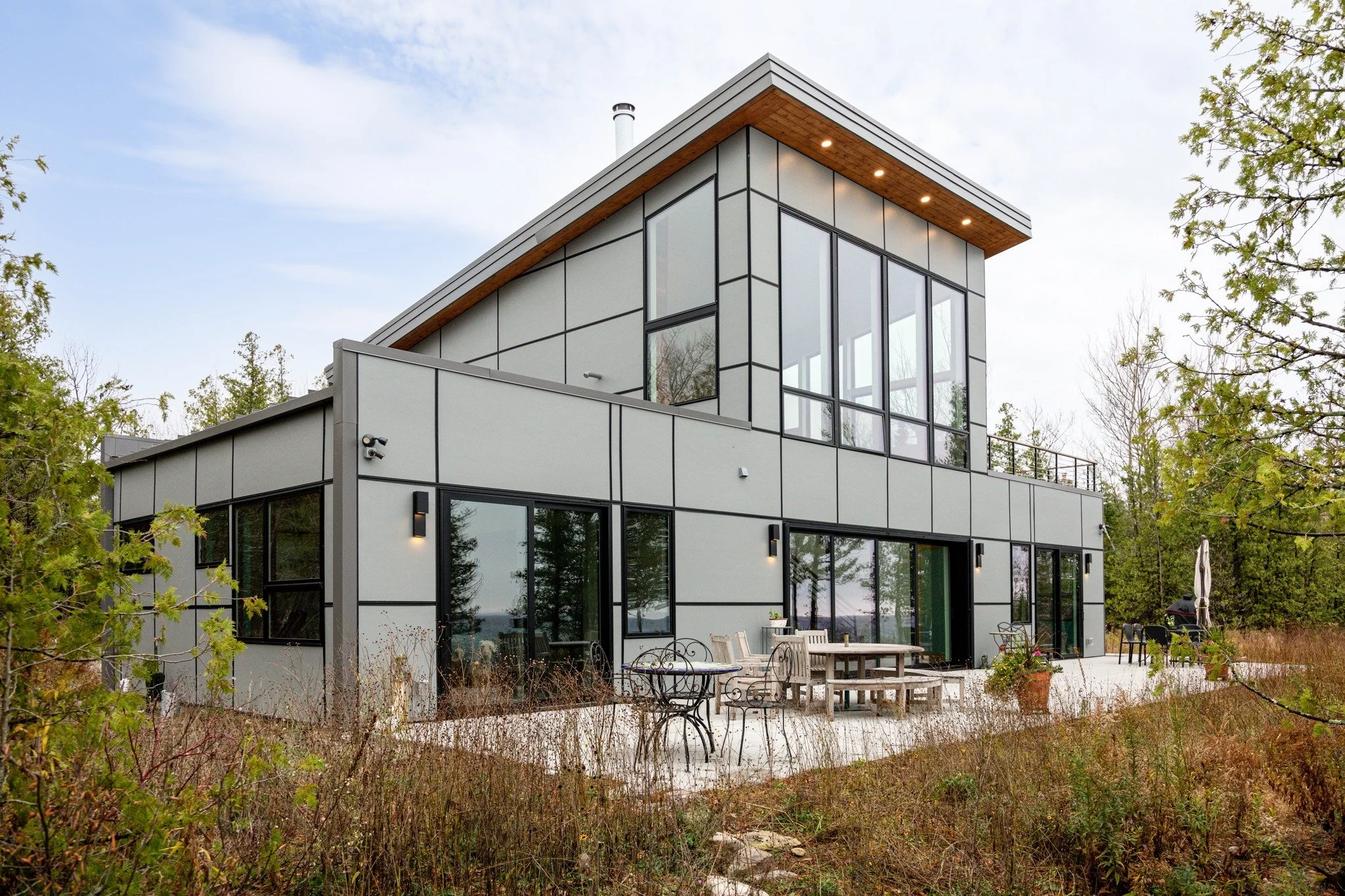 Modern lake home with gray and black siding, sloping roofs, a screened porch, and an outdoor seating deck among trees.