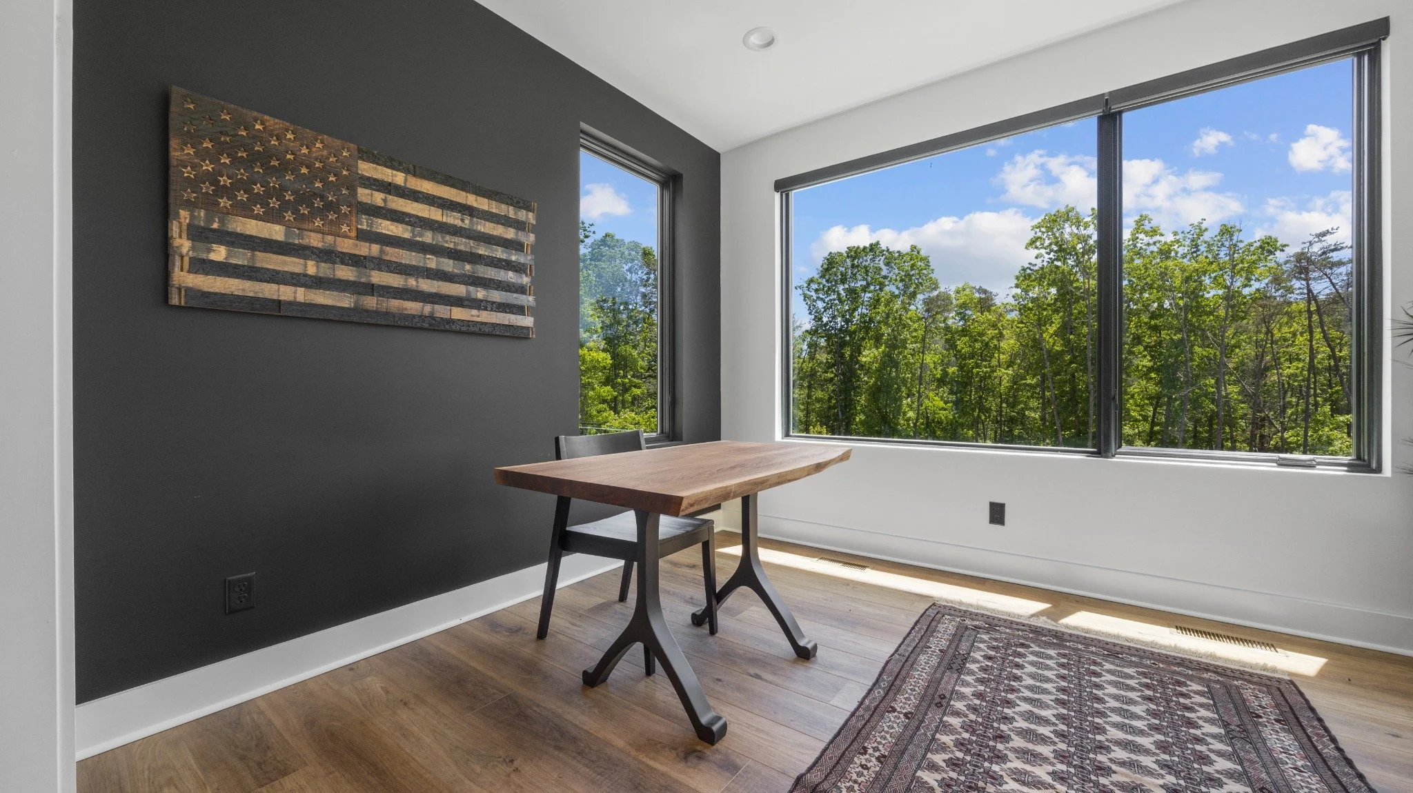 Custom home designer project featuring a wood desk, dark accent wall, wood flag artwork, and large windows overlooking trees.
