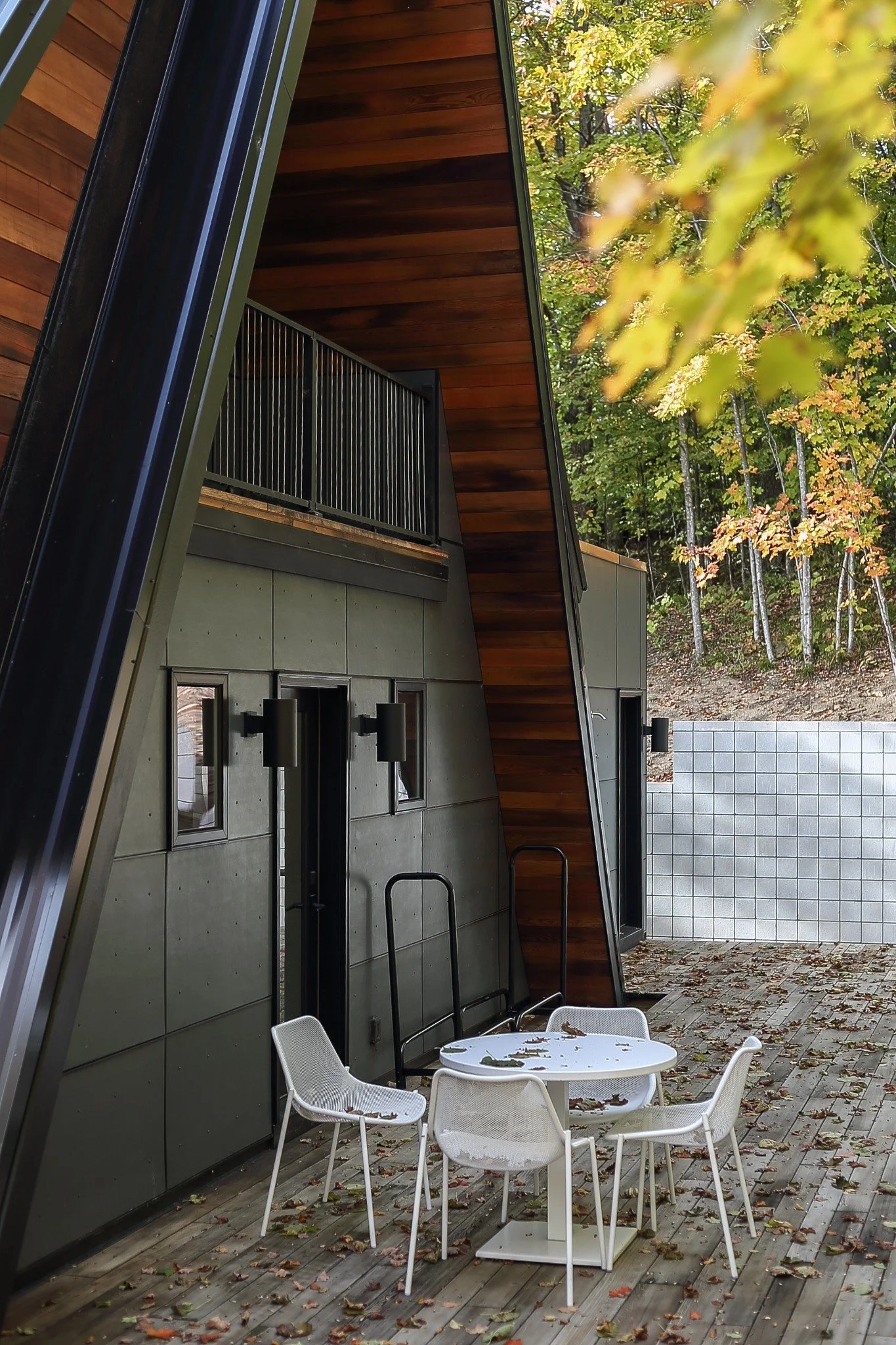 Lakefront home design featuring a dark A-frame exterior, triangular upper windows, and a raised deck surrounded by autumn trees