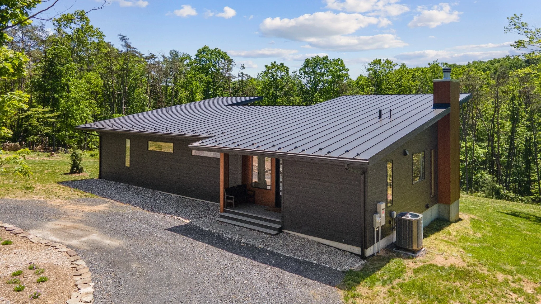 Mountain home architect project featuring a broad metal roof, dark siding, a wood-lined recessed entry, and a gravel driveway seen from above.