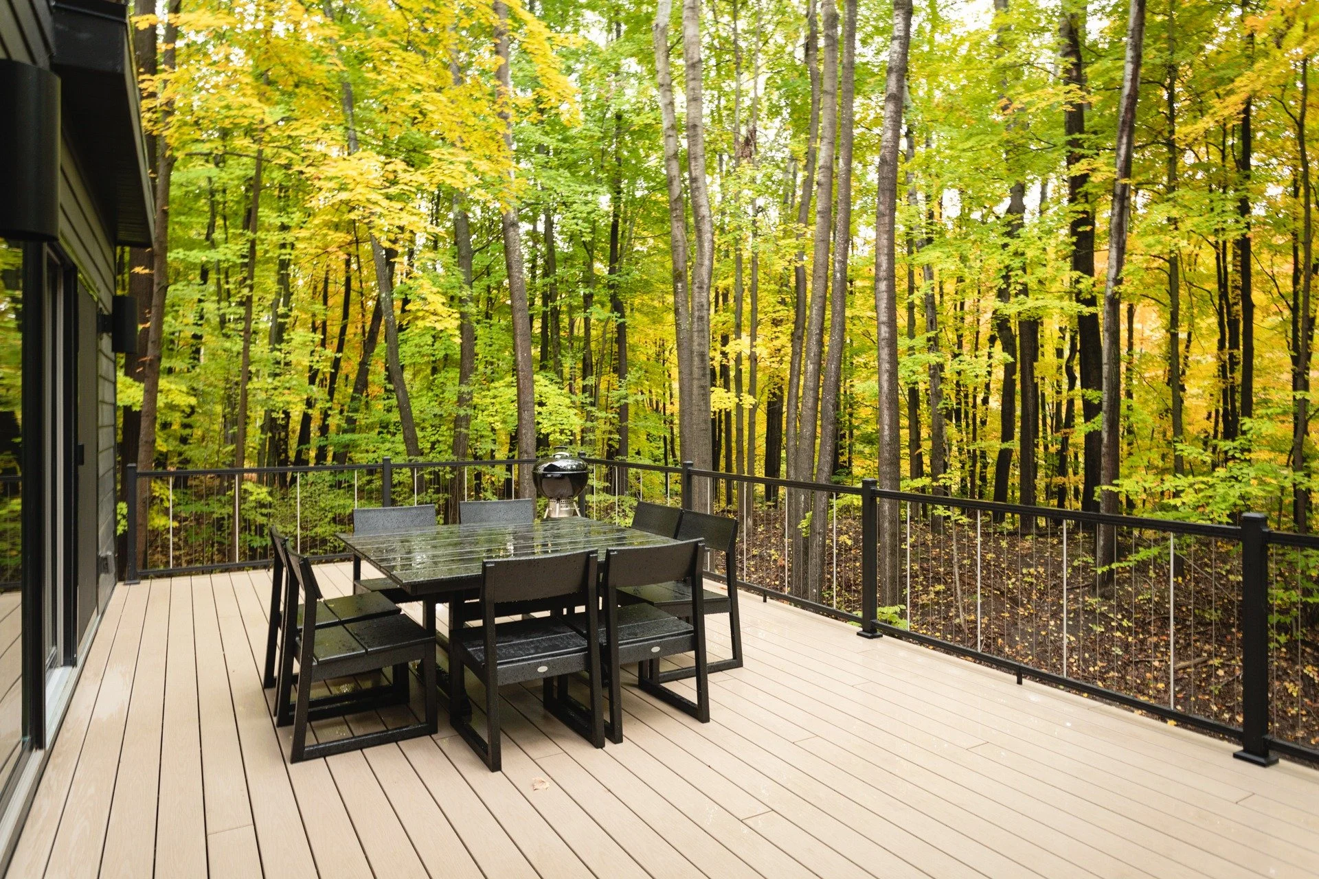 Custom mountain home designer project featuring a spacious deck, black dining furniture, a charcoal grill, and metal railings overlooking autumn woodland.
