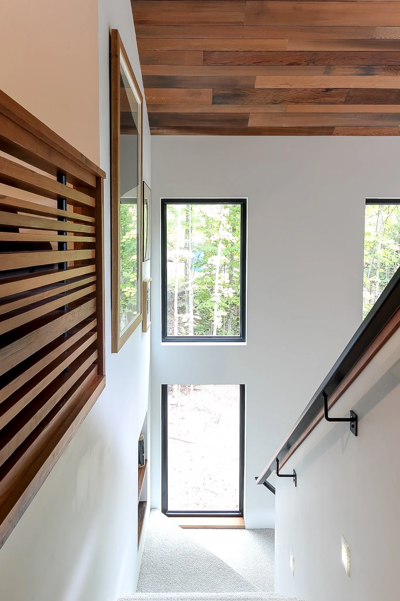  Lakeside house design with a bright stairwell, tall black-framed windows, a wood-slat railing, and a wood ceiling.
