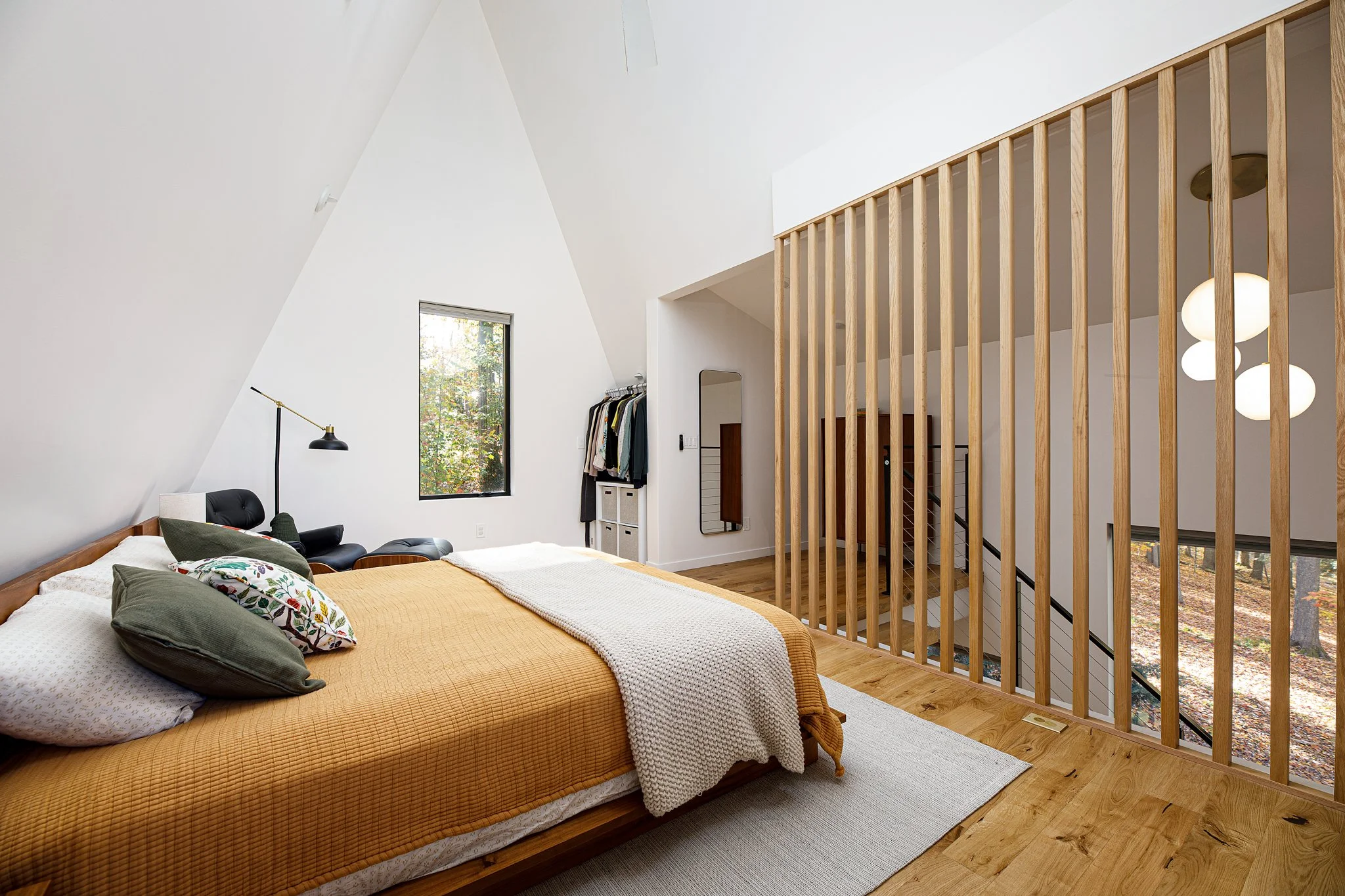 Bedroom with sloped ceiling, large window, yellow bedspread, throw blanket, headboard, pillows, black chaise lounge, clothing rack, mirror, hardwood floor, wooden staircase, and hanging white light fixtures.