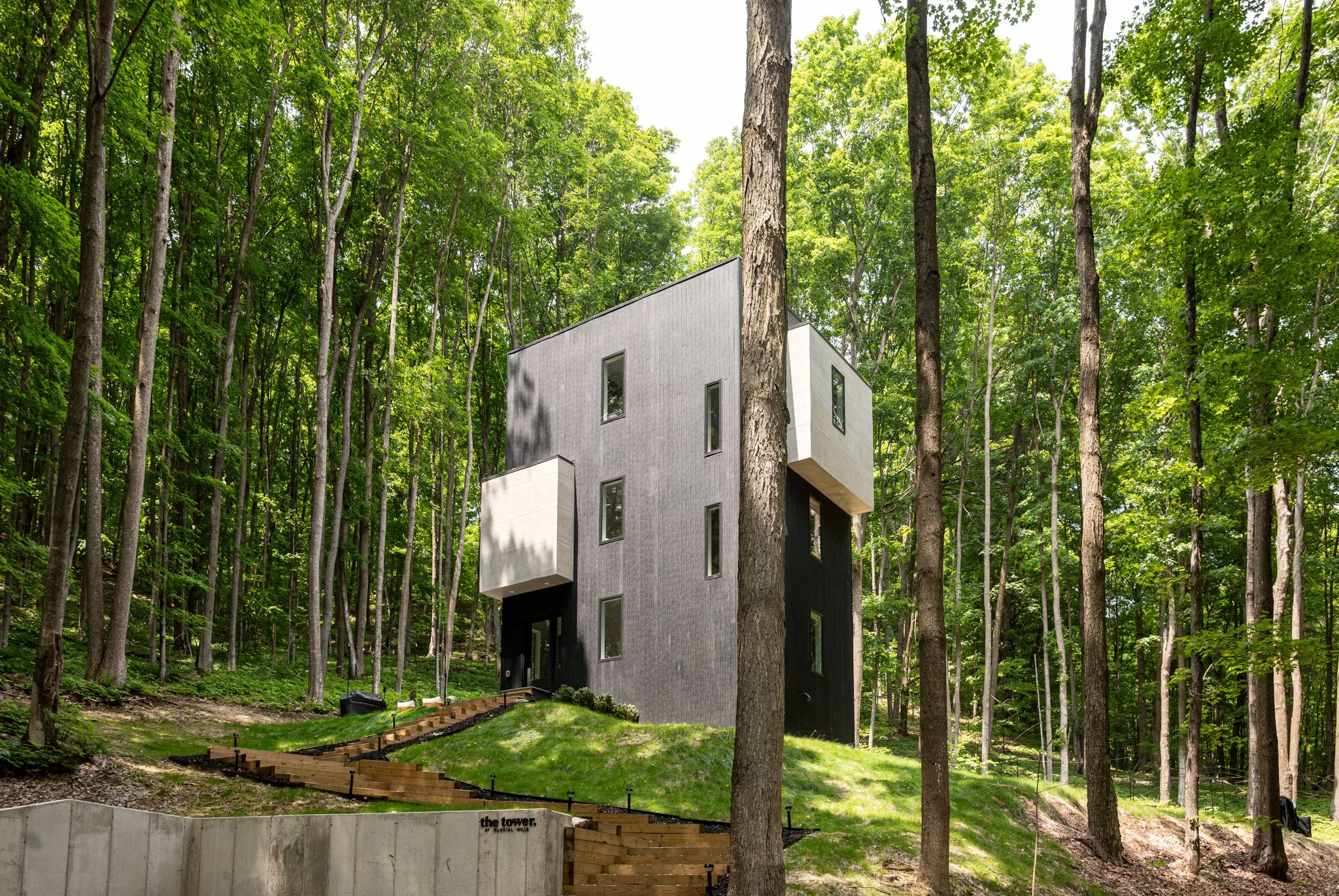 Modern house with gray and black exterior, surrounded by tall trees in a forest setting, with a wooden pathway leading up to the entrance.