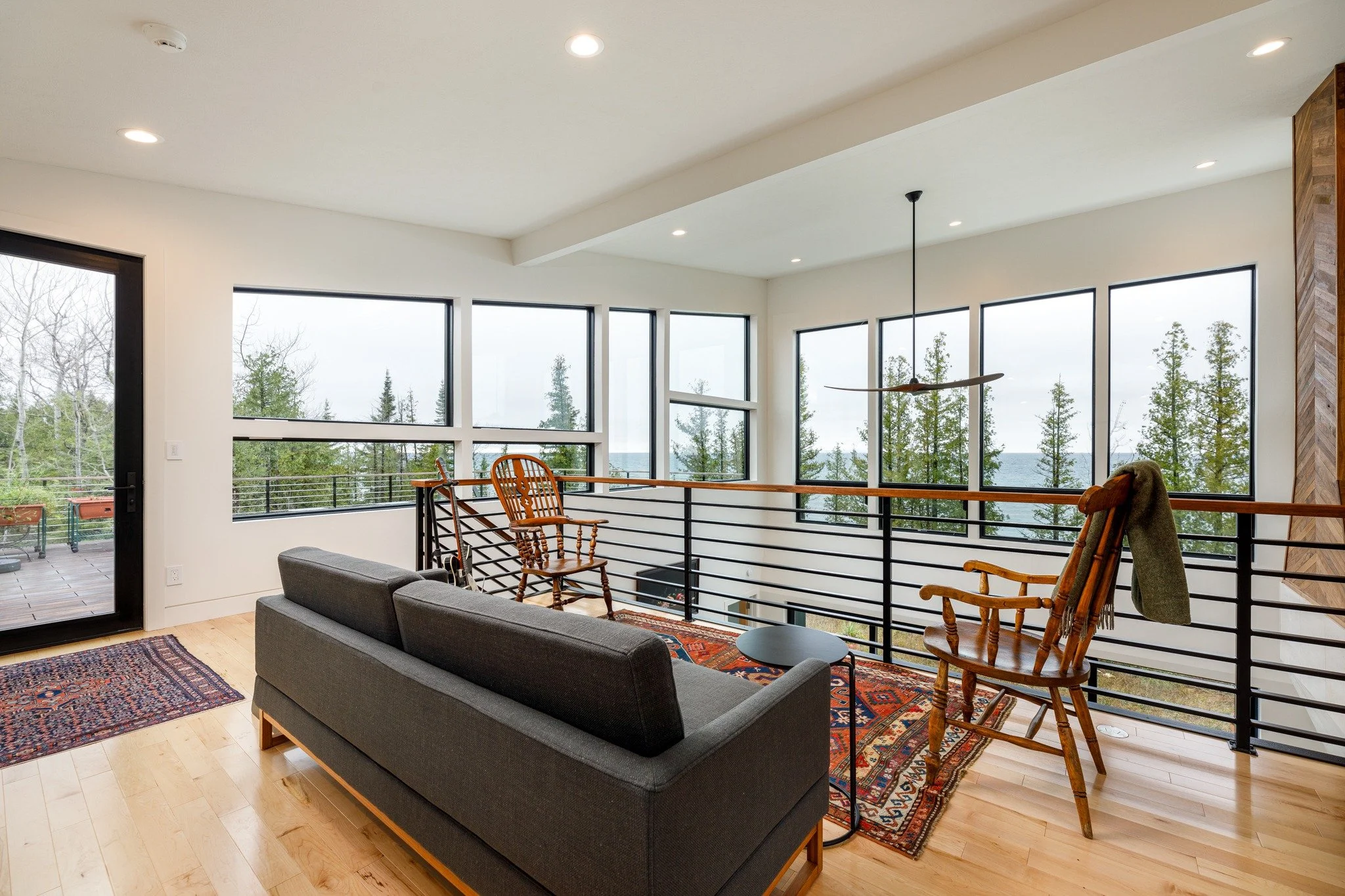 Lakefront home design with a loft sitting area, gray sofa, wooden rocking chairs, black railings, and water views.
