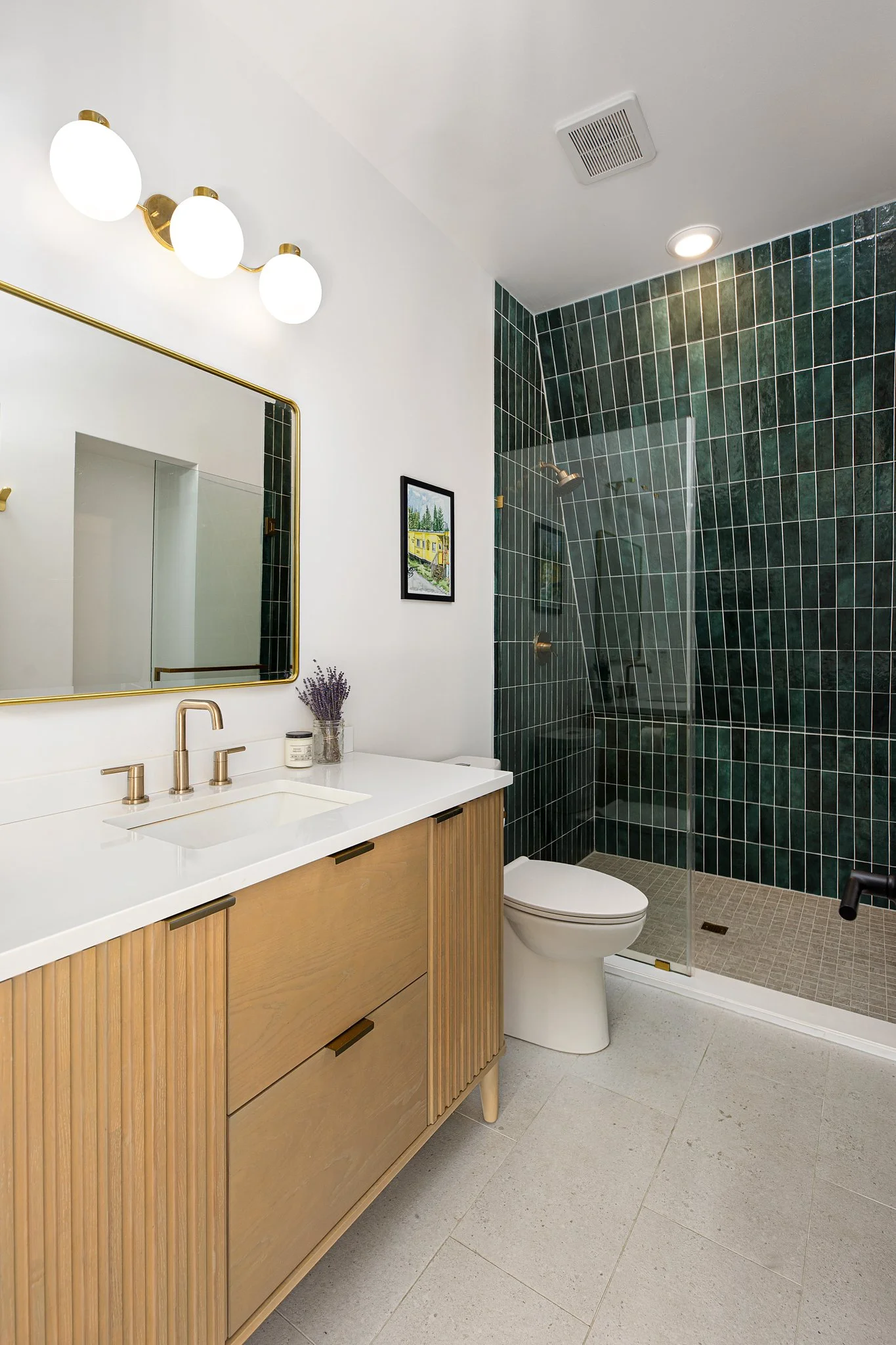Modern bathroom with wooden vanity, white countertop, and round white sink. Above the vanity is a large mirror with a gold frame. To the right, a walk-in shower with dark green tiles and a glass door. A small framed picture hangs on the wall, and a p